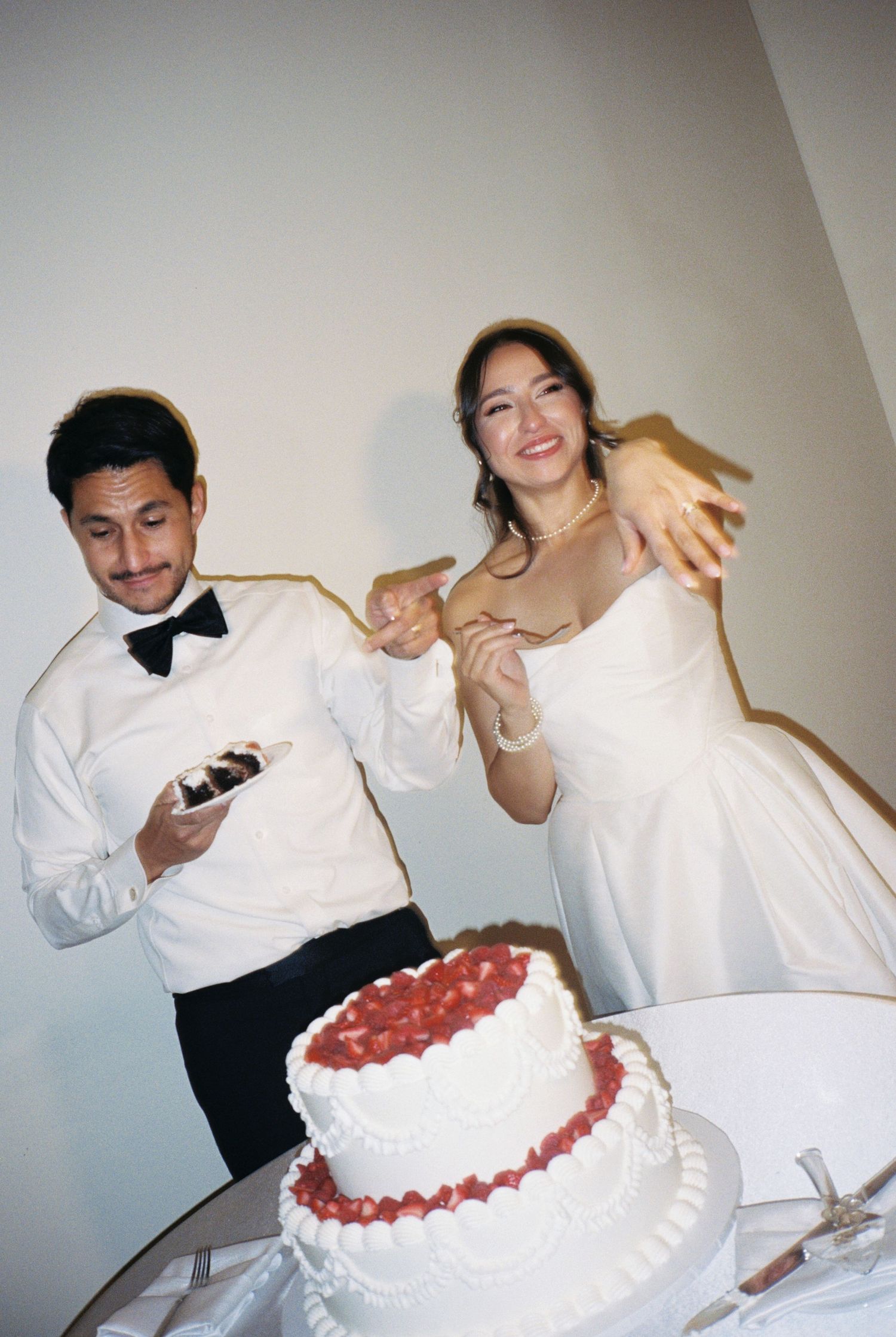 Bride and groom laughing while cutting a two-tier white wedding cake decorated with fresh strawberries.