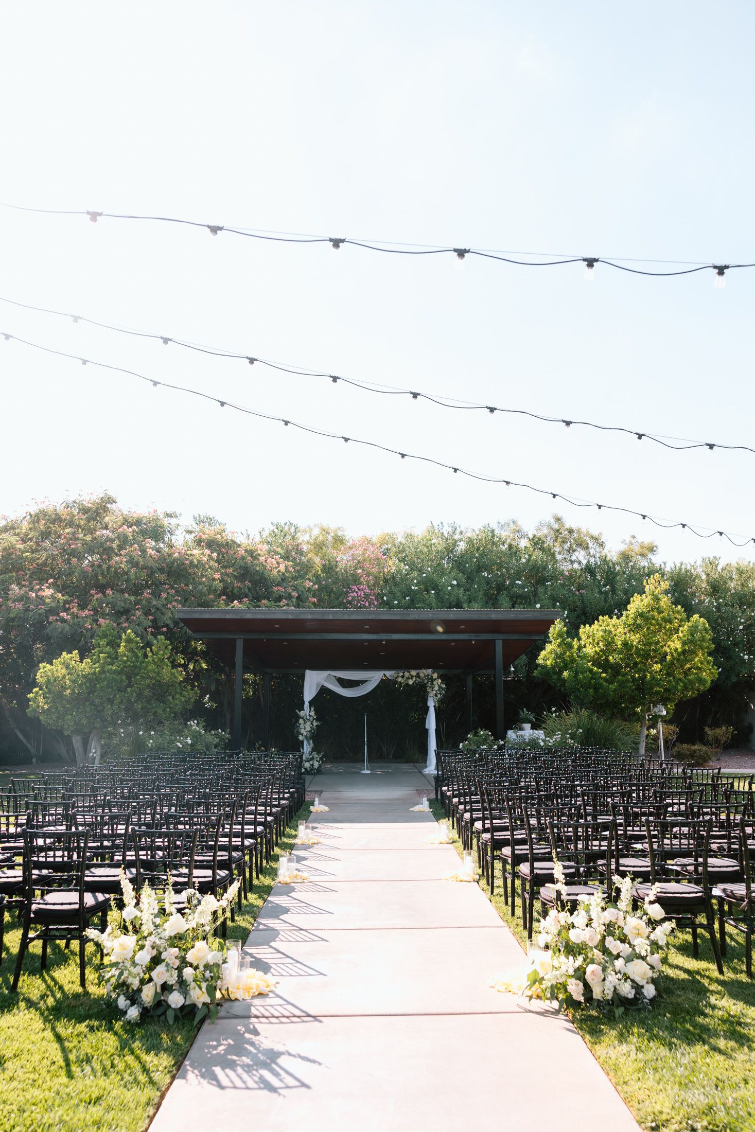 Outdoor wedding ceremony setup with rows of black chairs and string lights along a concrete pathway at sunset.