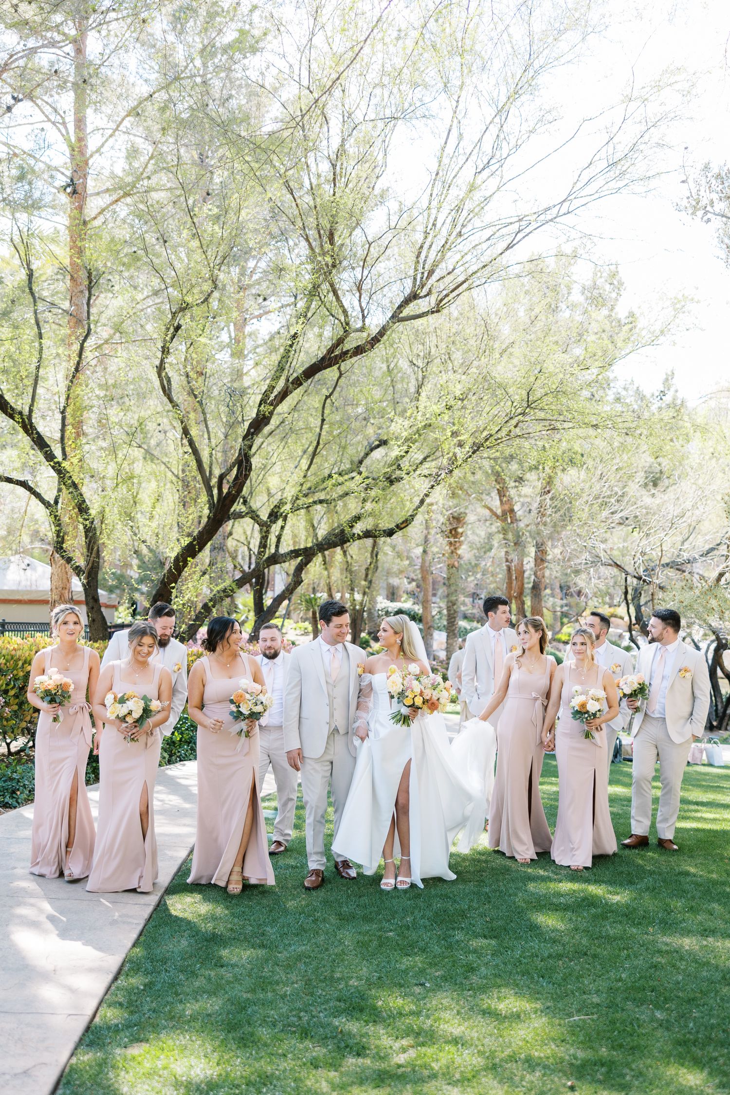 Wedding party walks outdoors, bridesmaids in blush gowns and groomsmen in tan suits with colorful bouquets.