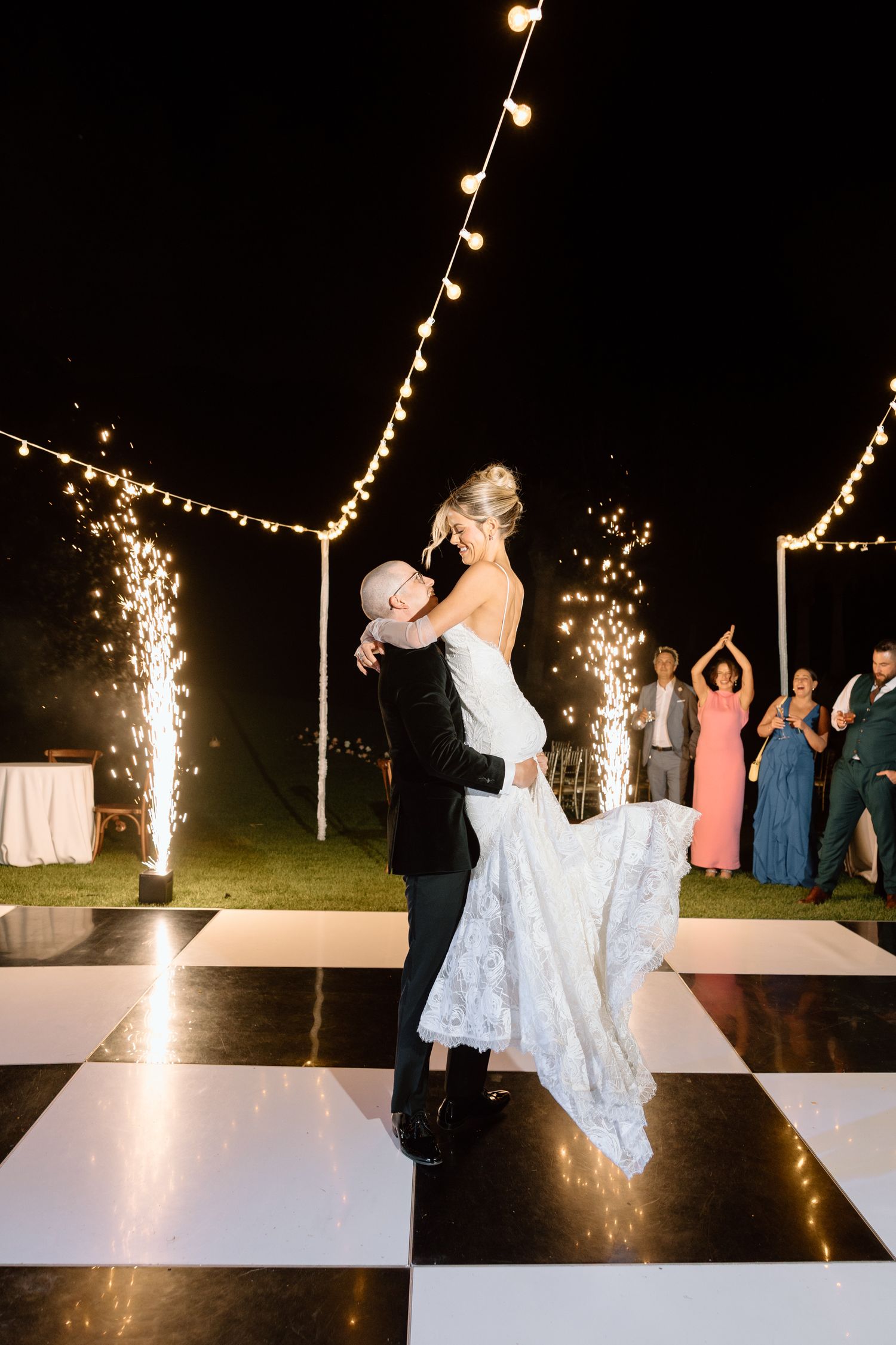 Groom lifts bride on checkered dance floor under string lights at outdoor wedding reception at night.