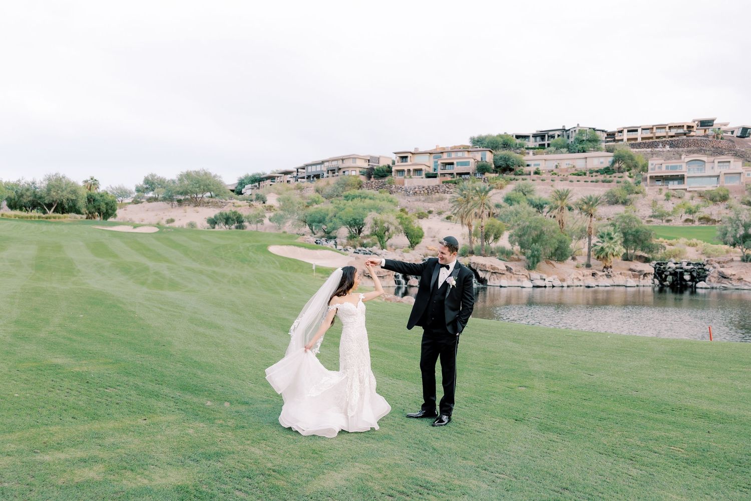 Bride and groom dancing on lush green golf course with scenic lake and hillside venue in background.