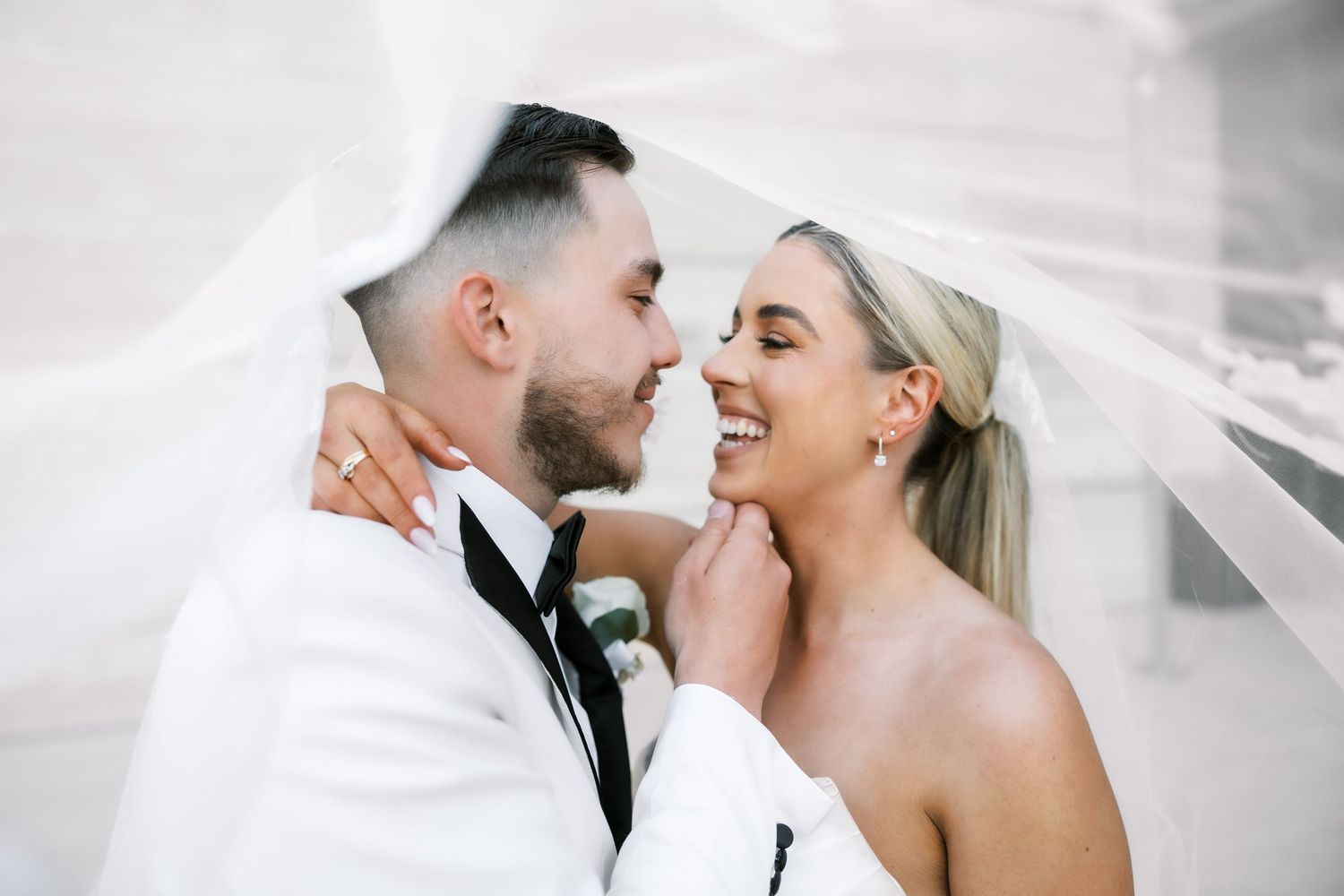 Bride and groom share a romantic moment under a flowing veil, smiling at each other on their wedding day.