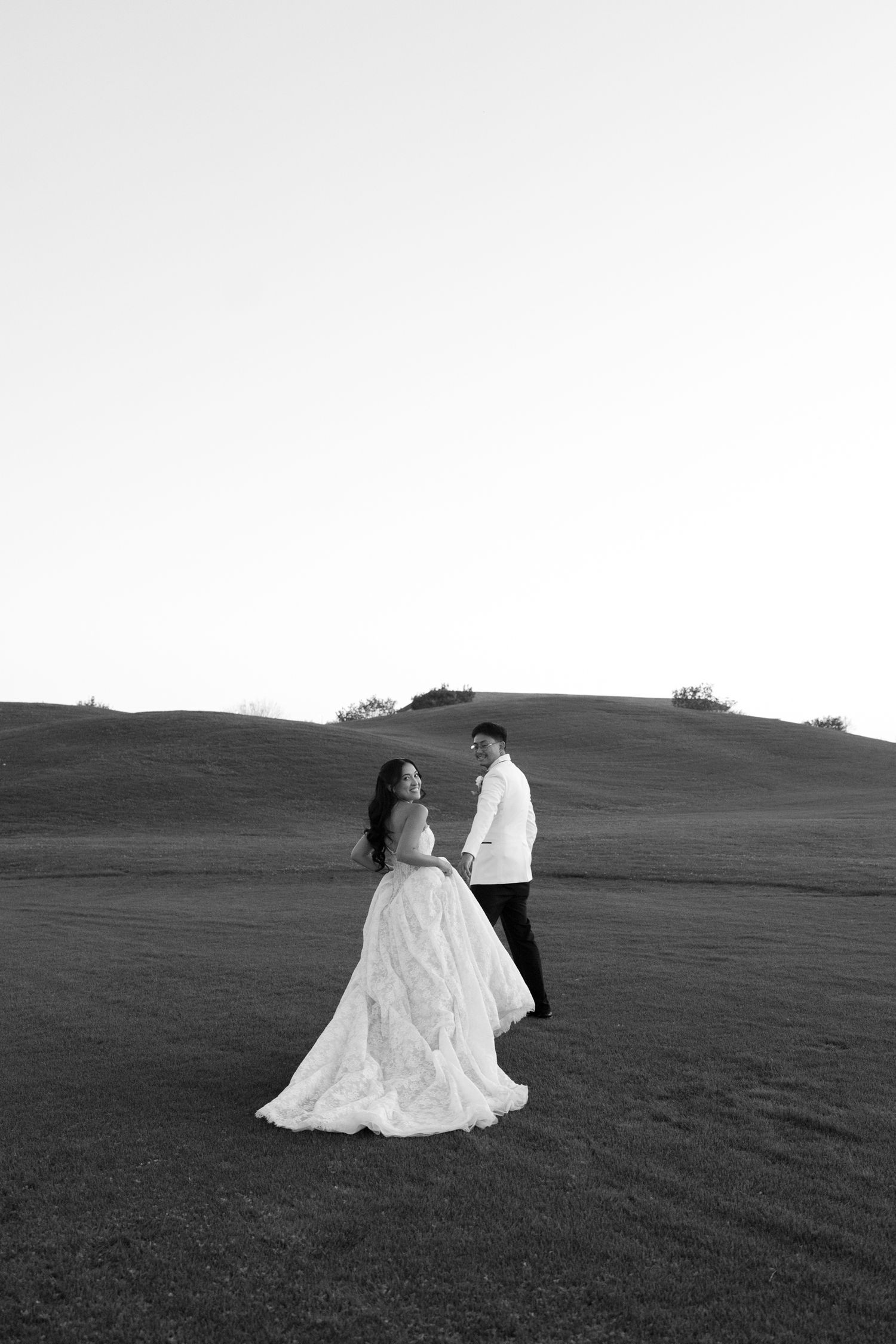 Black and white photo of a bride in a flowing lace gown and groom in a white jacket walking hand in hand across a grassy hillside.