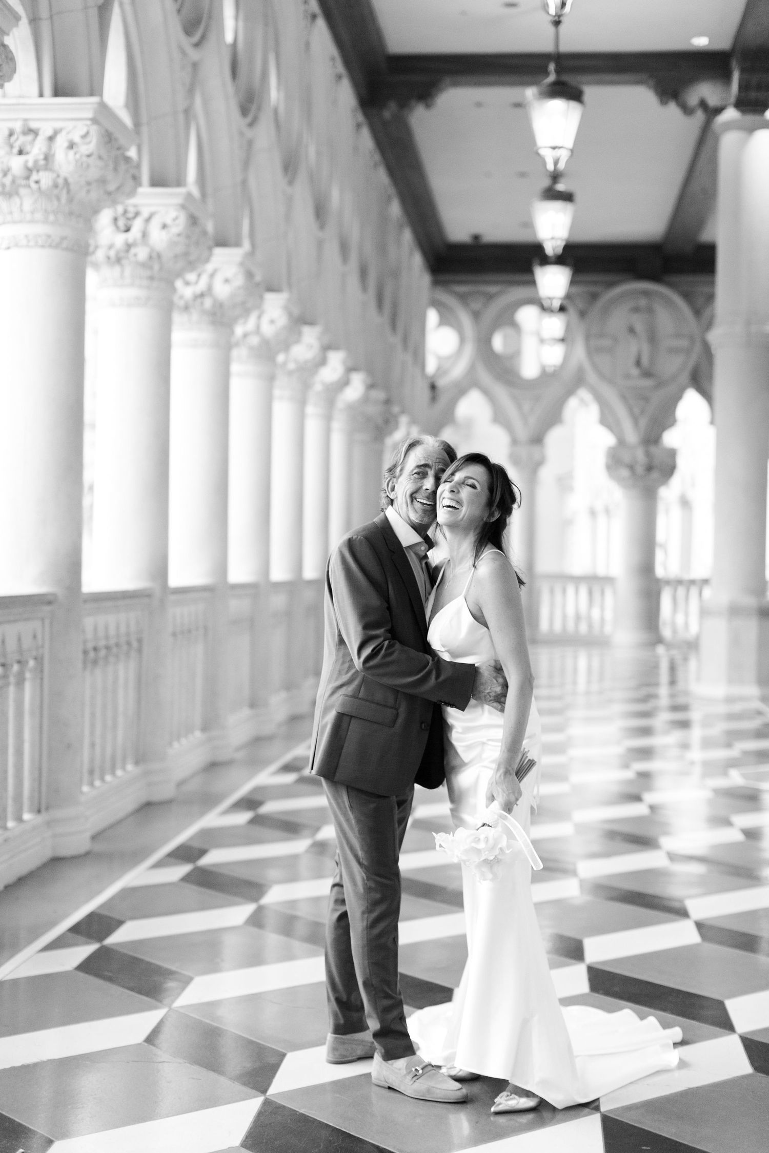 Black and white photo of a couple in wedding attire embracing in an elegant columned hallway with checkered marble floors.