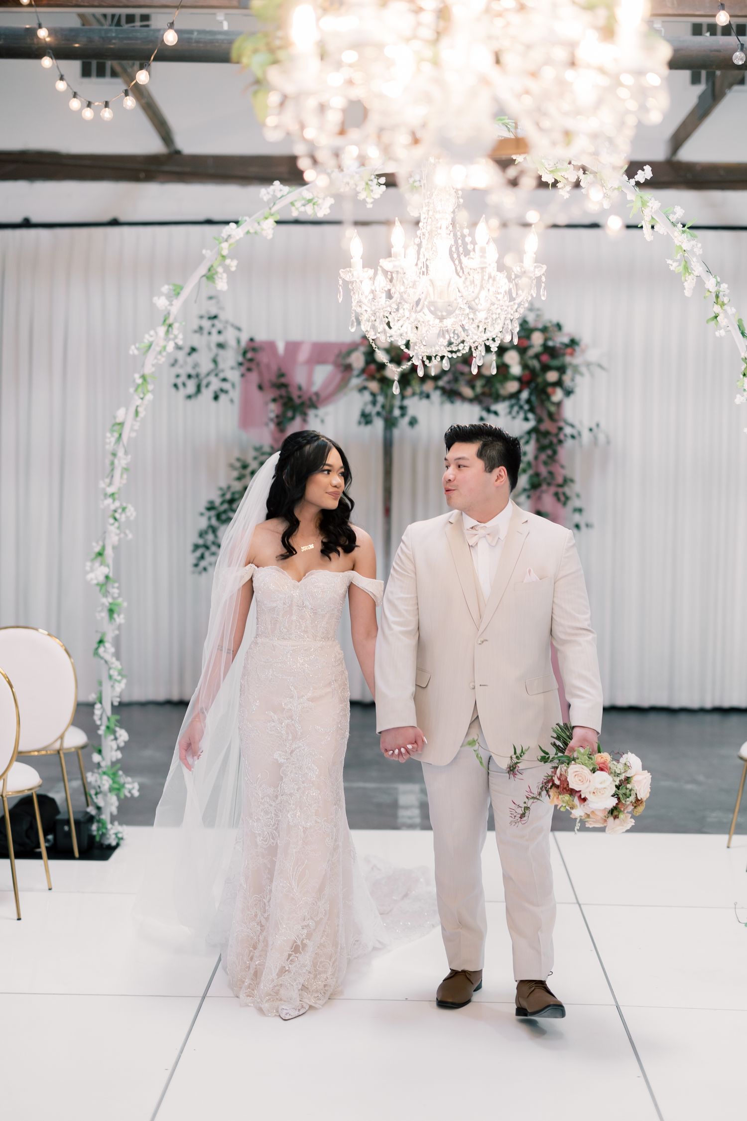 Wedding couple dancing in a bright white reception venue under sparkling crystal chandeliers.