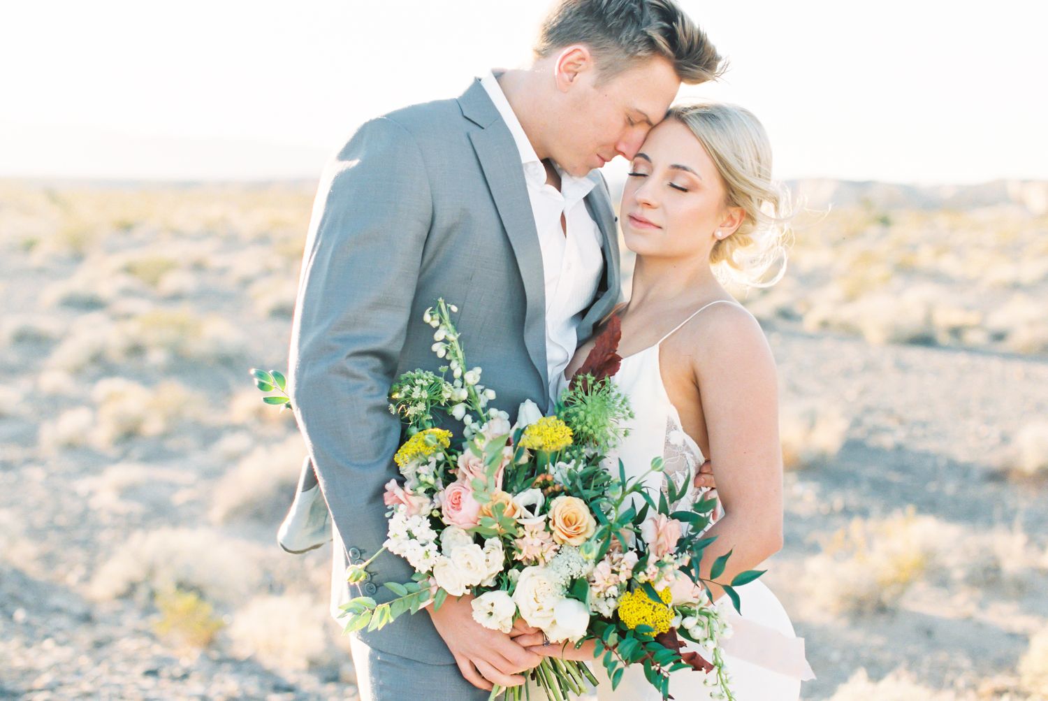 Romantic couple embracing in desert landscape, groom in gray suit holding lush floral bouquet with bride in white gown.