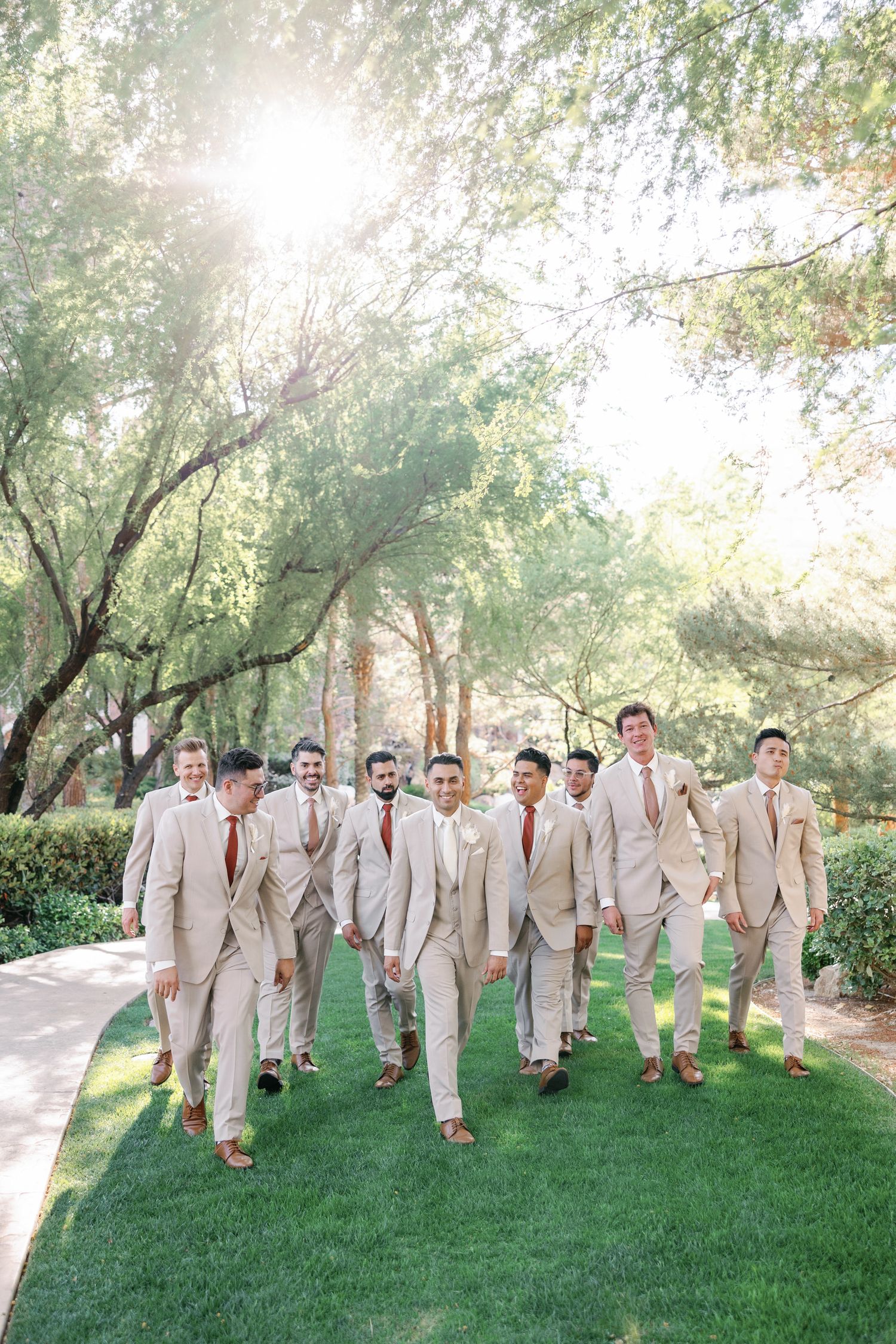 Groomsmen in tan suits with rust ties walk together on a lush green lawn surrounded by trees.