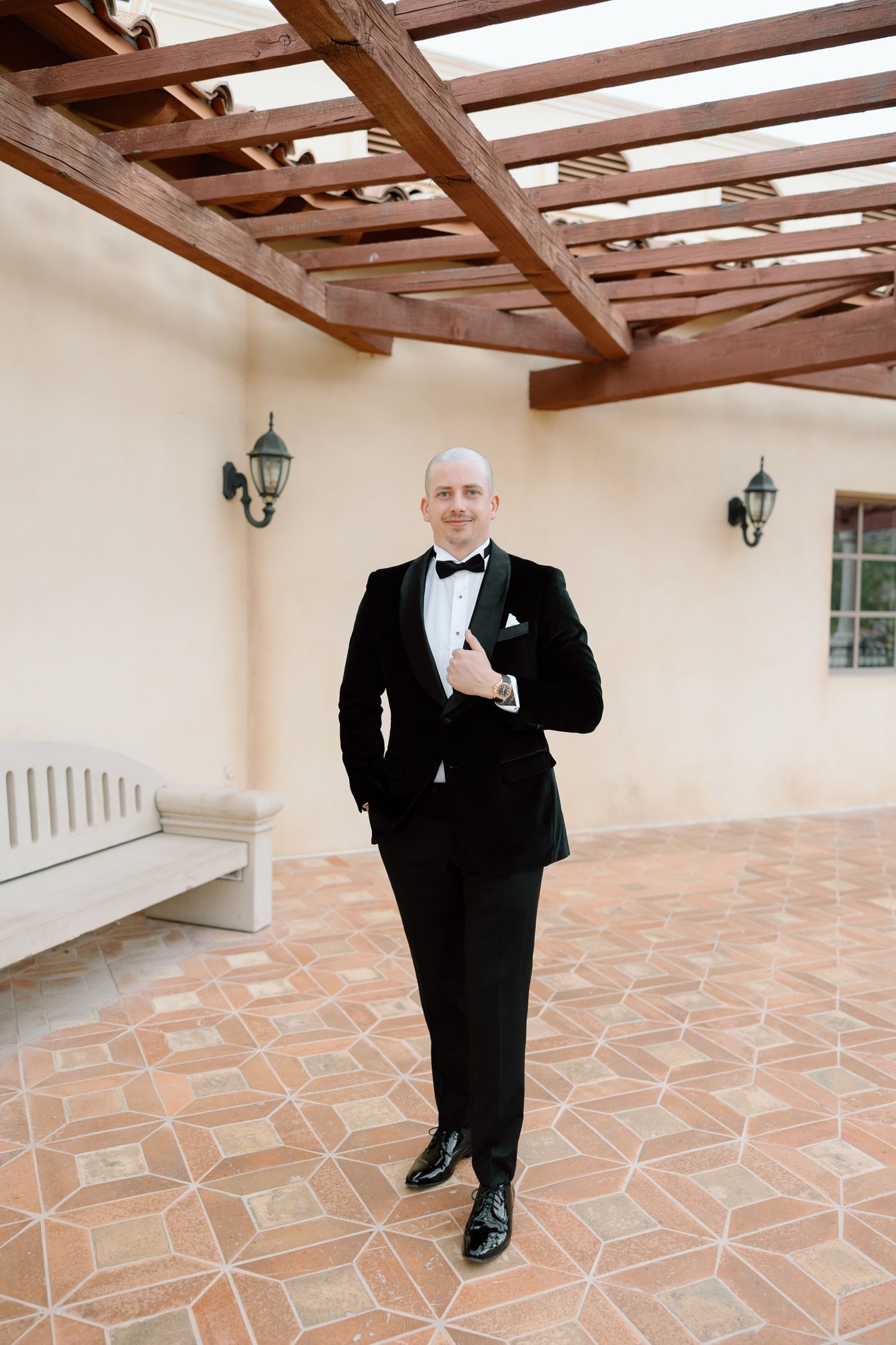 A dapper groom in a black tuxedo with bow tie stands confidently on a terracotta patio beneath a wooden pergola.