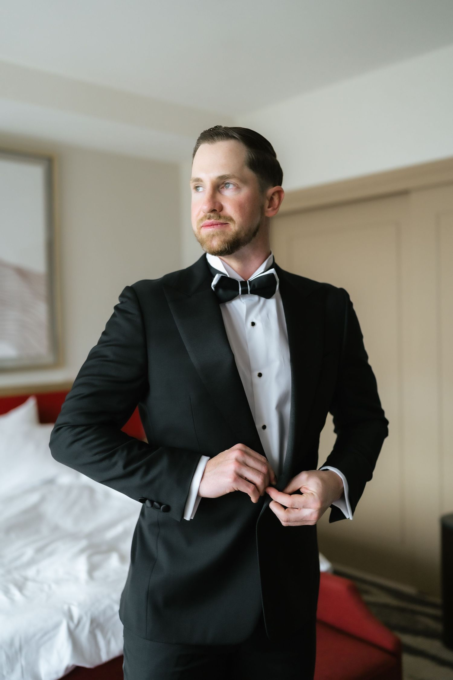 Groom in black tuxedo with bow tie buttoning jacket in hotel room before wedding ceremony.