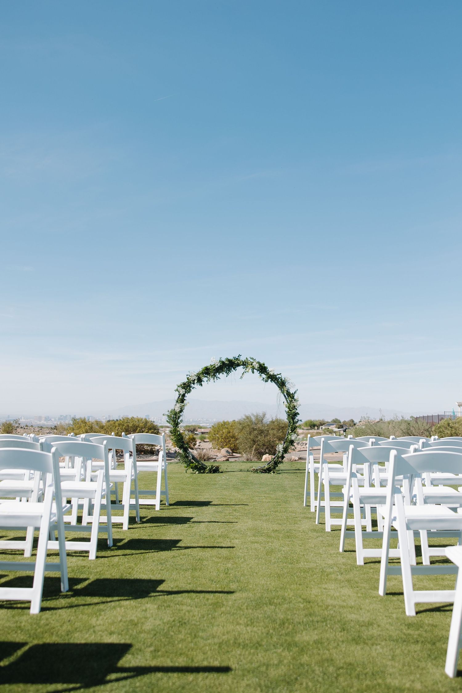 Outdoor wedding ceremony setup with white chairs lining a green aisle leading to a circular floral arch under a clear blue sky.