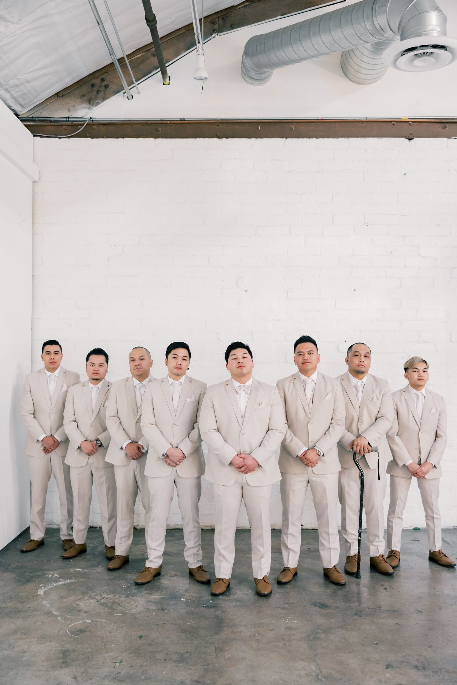 Groomsmen in light beige suits stand in a row against a white industrial wall.