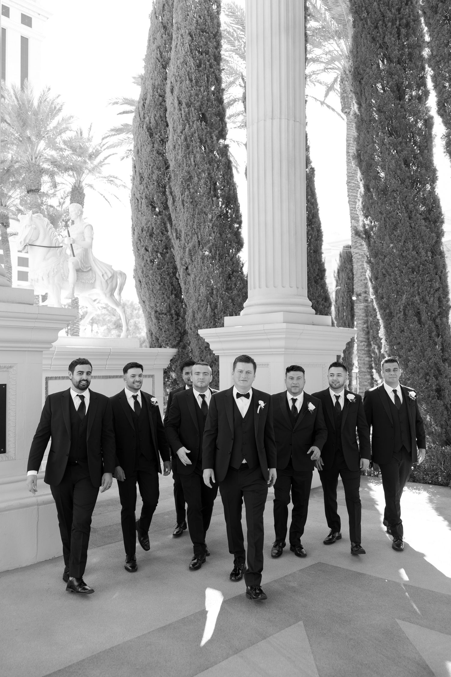 A group of groomsmen in black suits walk together along a path with white columns and cypress trees in black and white.