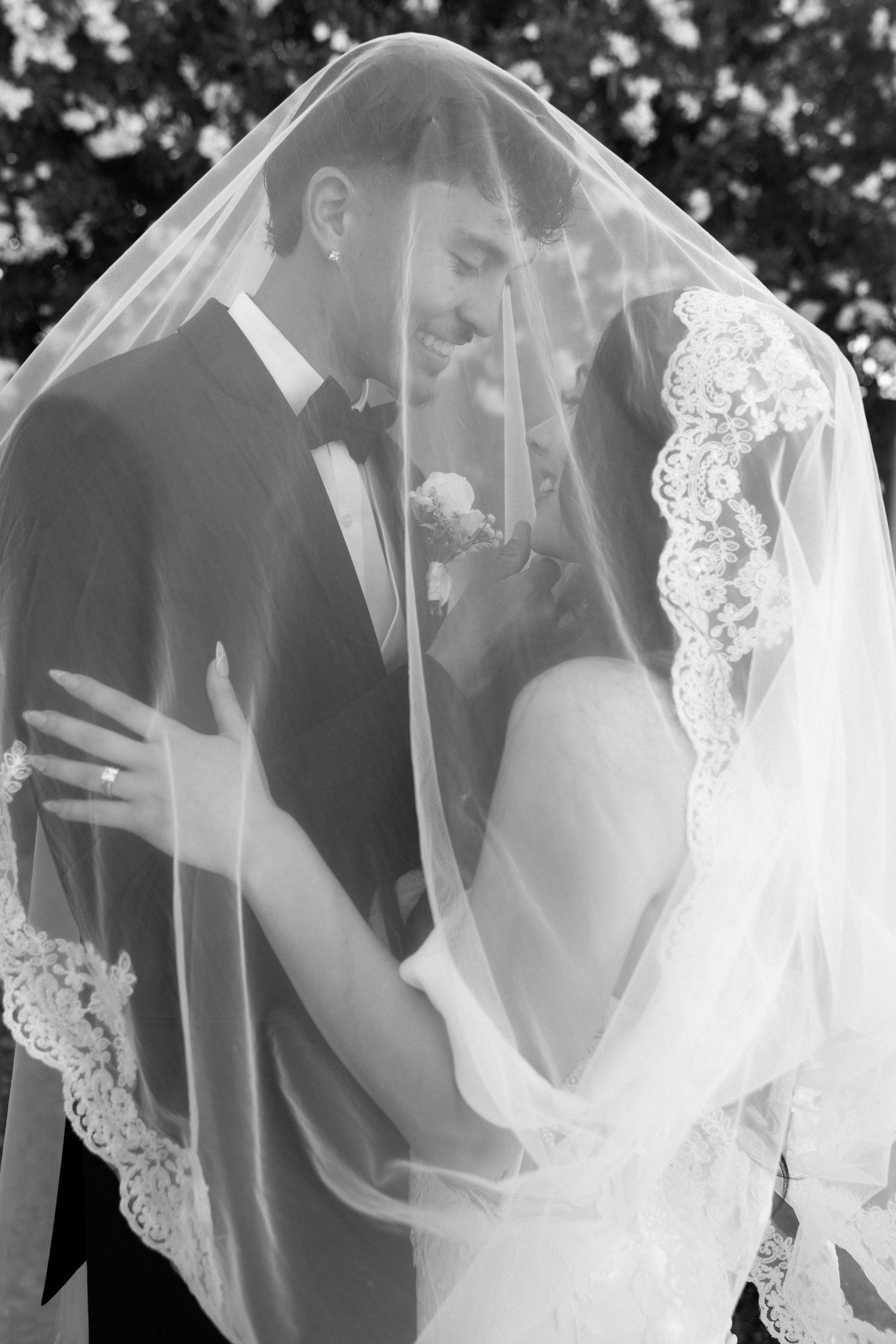 Black and white photo of bride and groom sharing intimate moment under lace wedding veil.