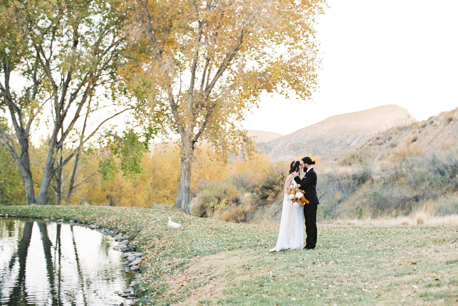 Couple shares romantic kiss by a serene pond surrounded by autumn trees and rolling hills on their wedding day.