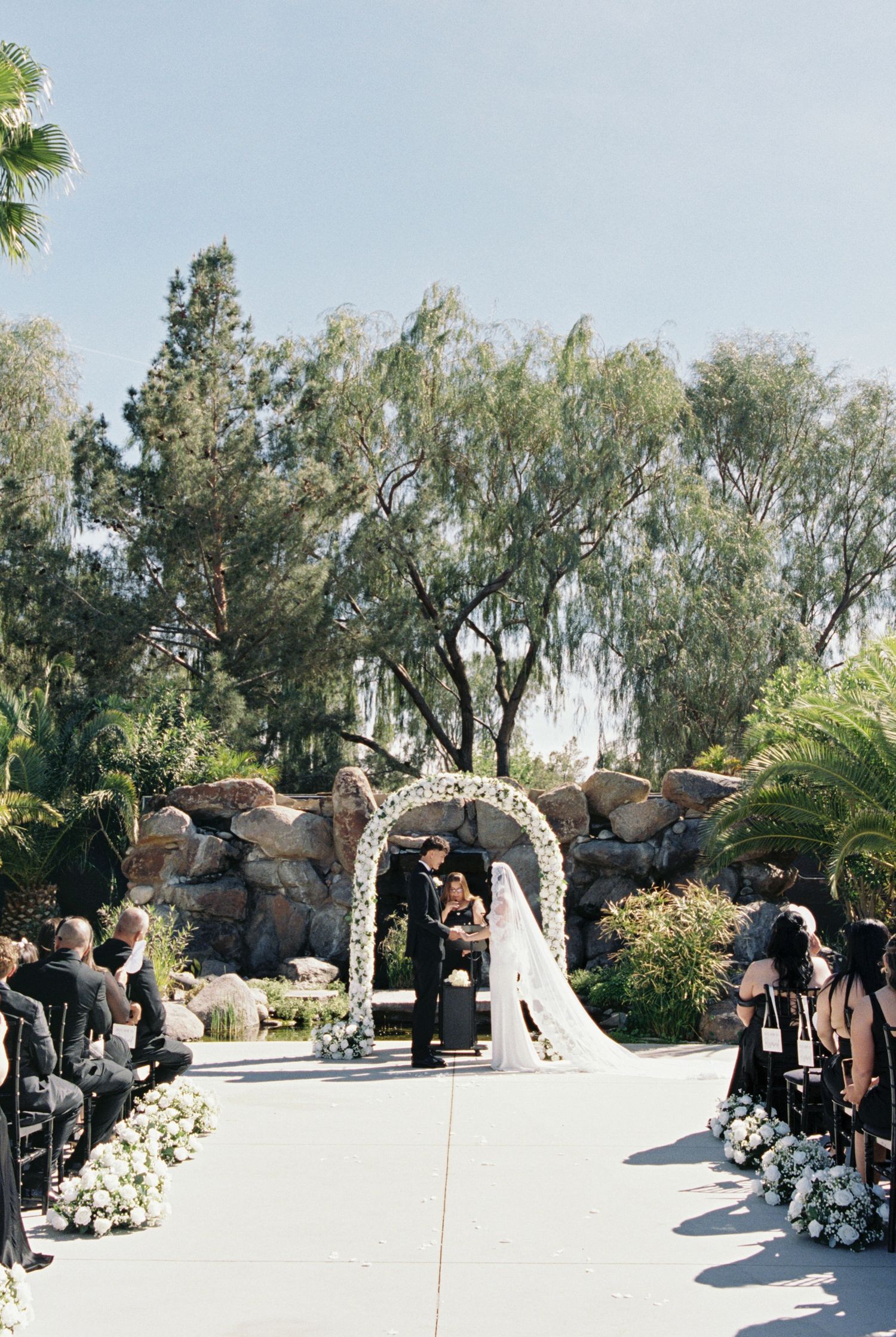 Outdoor wedding ceremony with floral arch, bride and groom at altar, white flowers lining aisle, lush trees and rock backdrop.