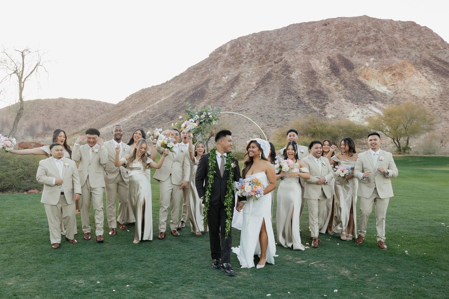 Wedding party in cream attire poses on golf course with desert mountain backdrop under circular floral arch.