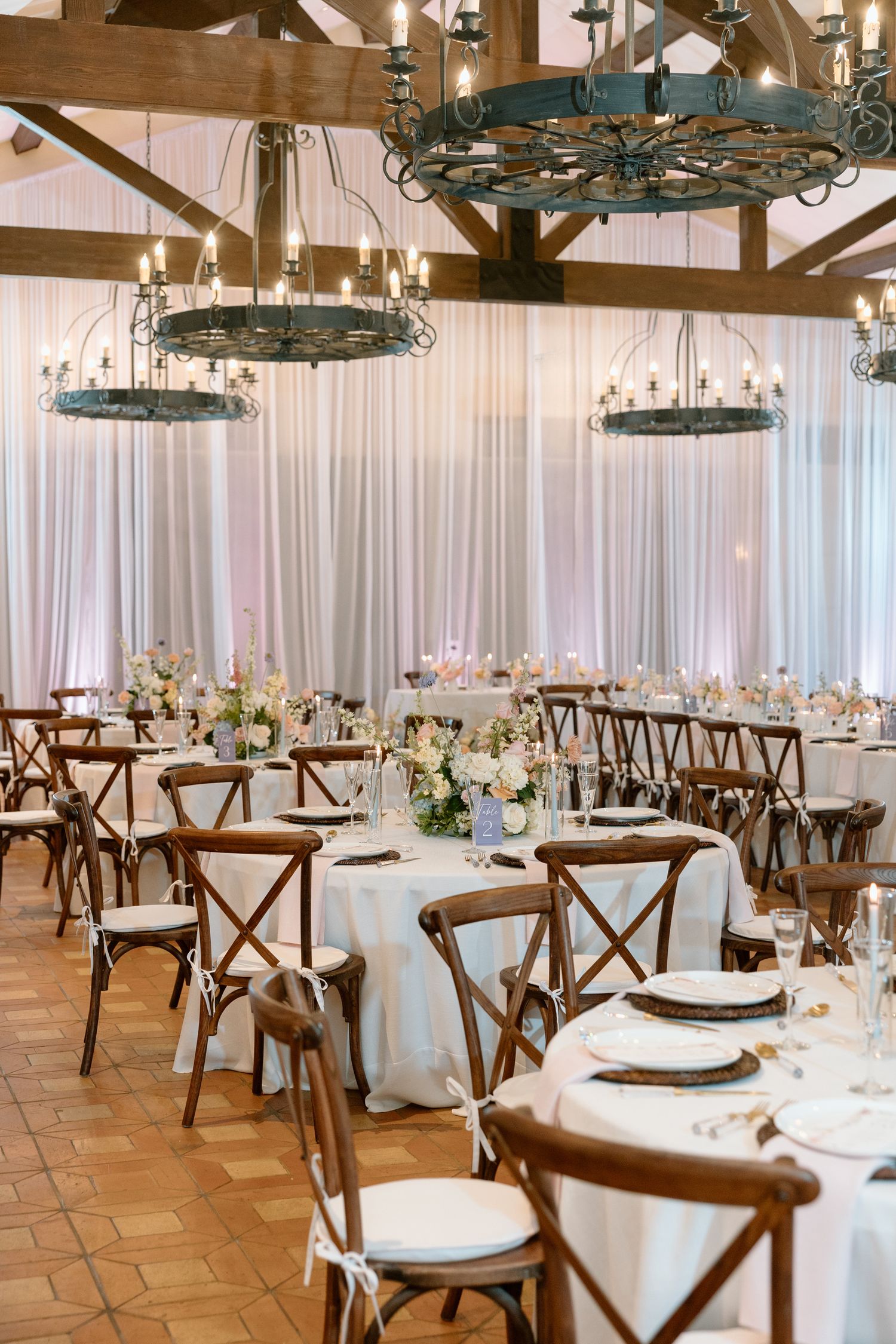 Elegant wedding reception hall with rustic wood chairs, round tables, white linens, and vintage wagon wheel chandeliers.