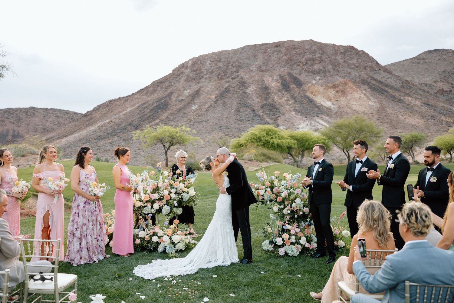 Outdoor desert wedding ceremony with bride and groom kissing, floral arch, bridesmaids in pink, groomsmen in black, mountain backdrop.
