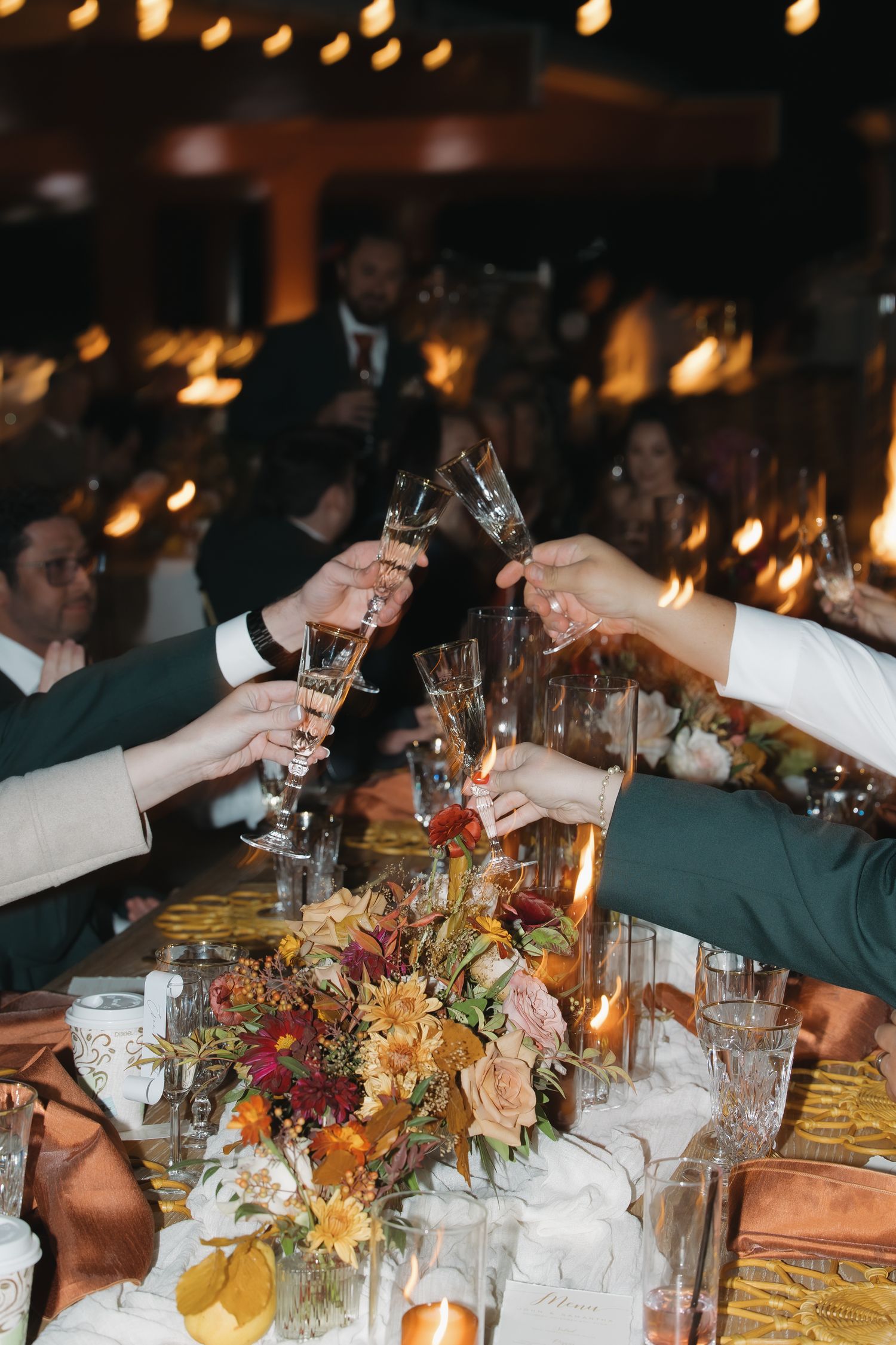 Wedding guests raise champagne glasses in a toast over a table with autumn floral centerpieces.