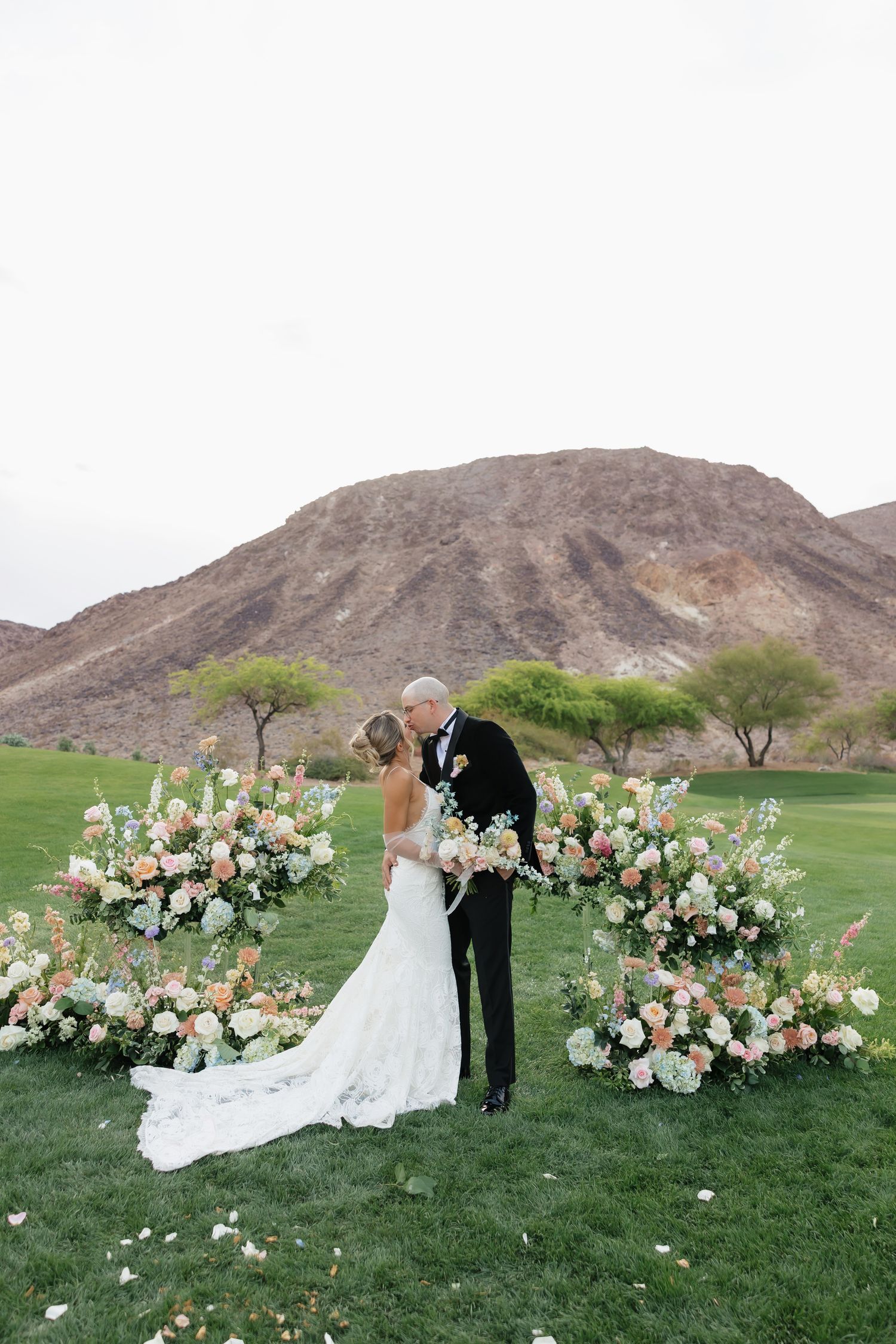 Bride and groom share a kiss at outdoor desert wedding ceremony with floral arrangements and mountain backdrop.