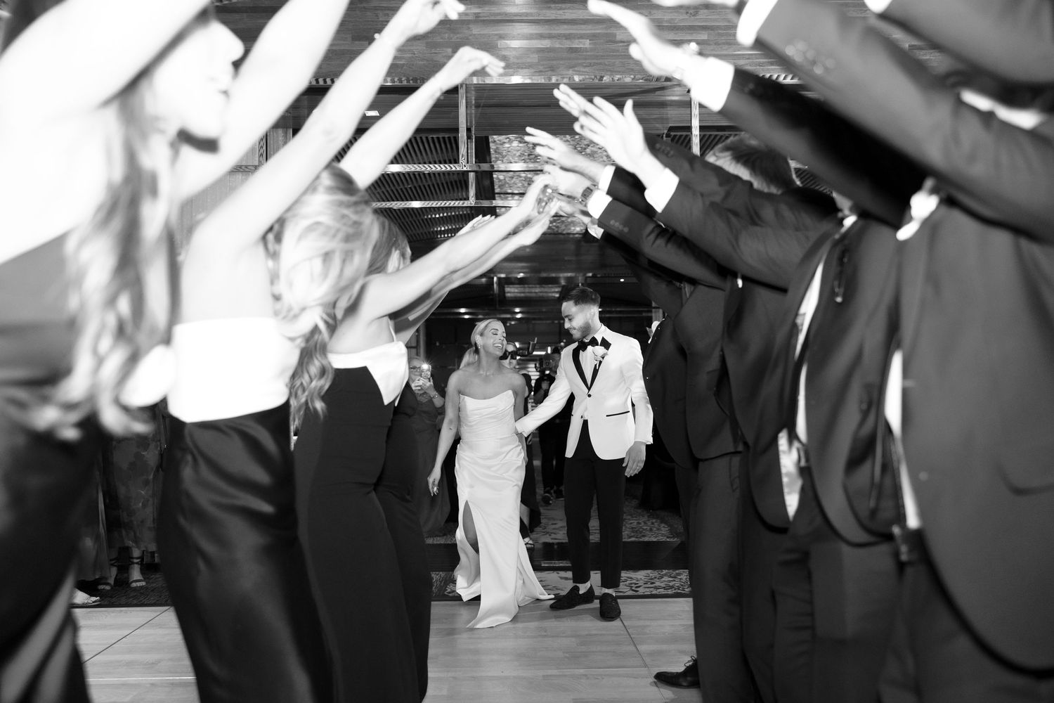Black and white photo of bride and groom walking through a tunnel of raised hands at their wedding reception.