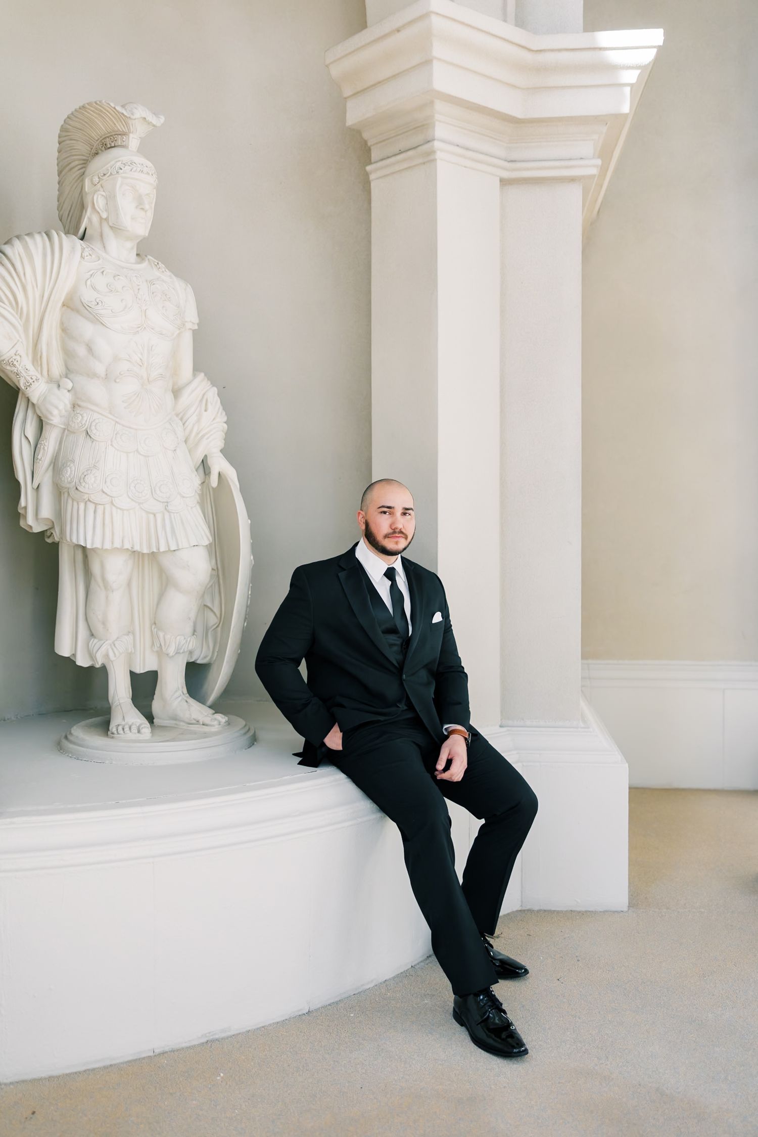 A bald man in a black suit sits beside a white Roman soldier statue next to an elegant column in a classical interior.