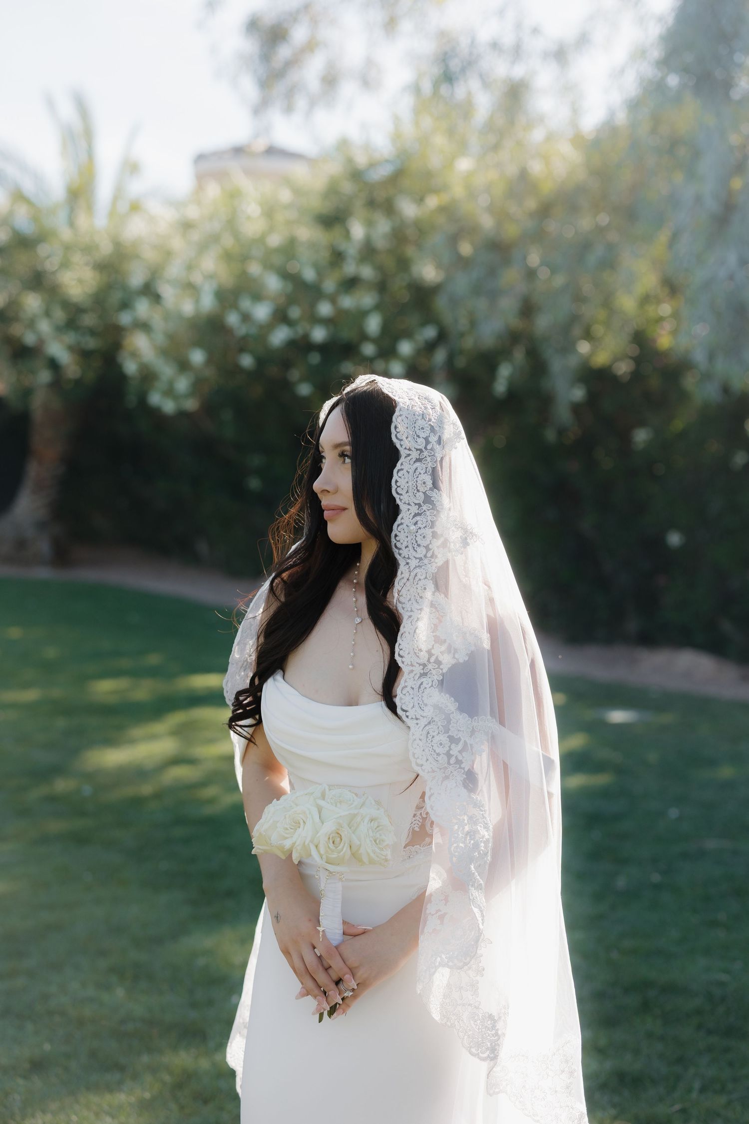 Elegant bride in white gown and lace veil holding bouquet outdoors in a lush garden.