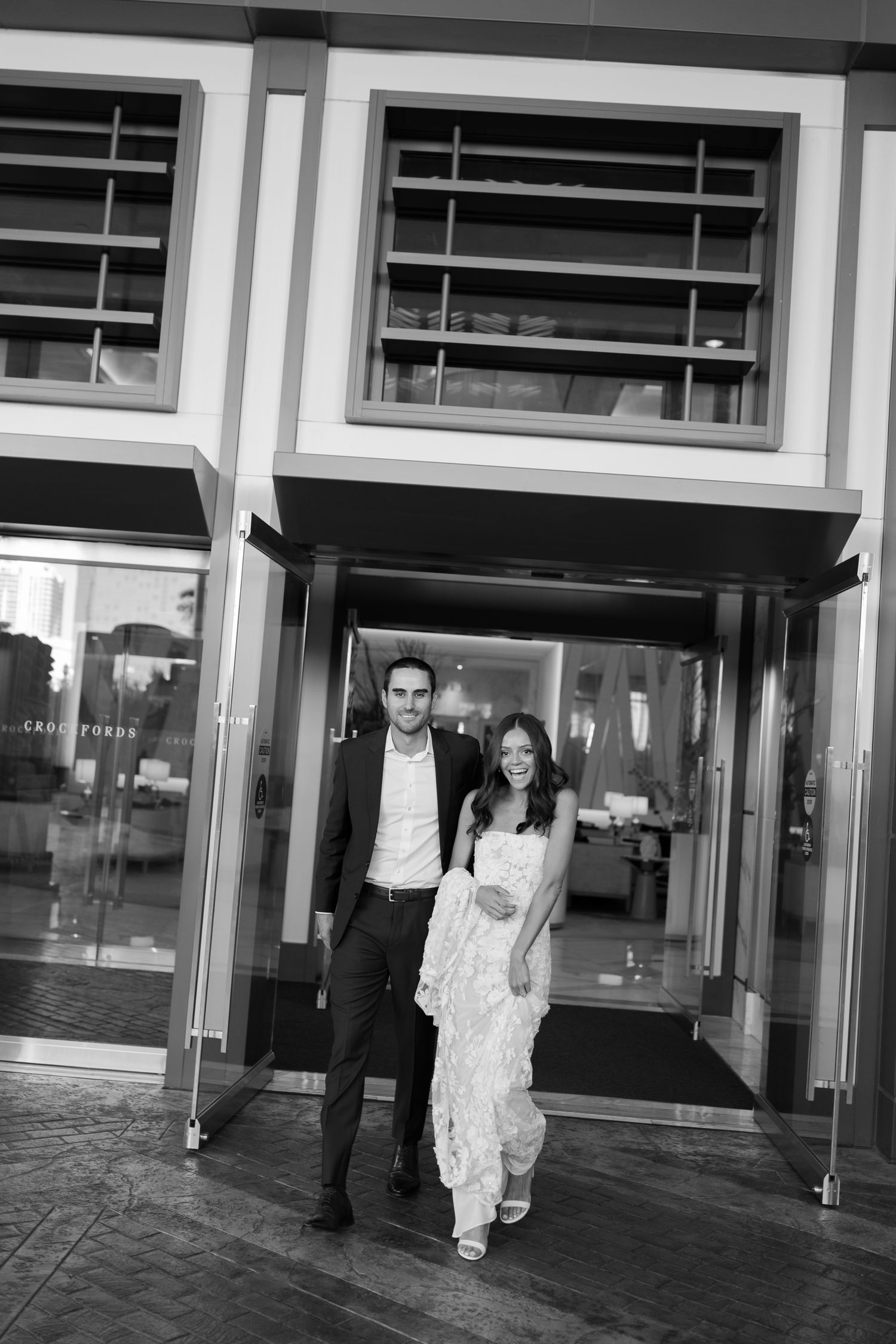 Black and white photo of an elegant couple exiting a modern building, woman in white dress, man in dark suit.