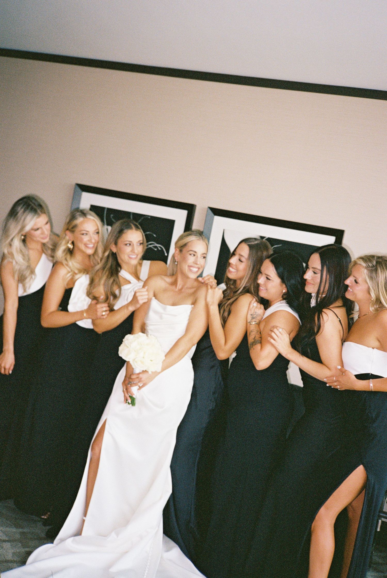 Bride in white gown holds bouquet surrounded by bridesmaids in black dresses, laughing together indoors.