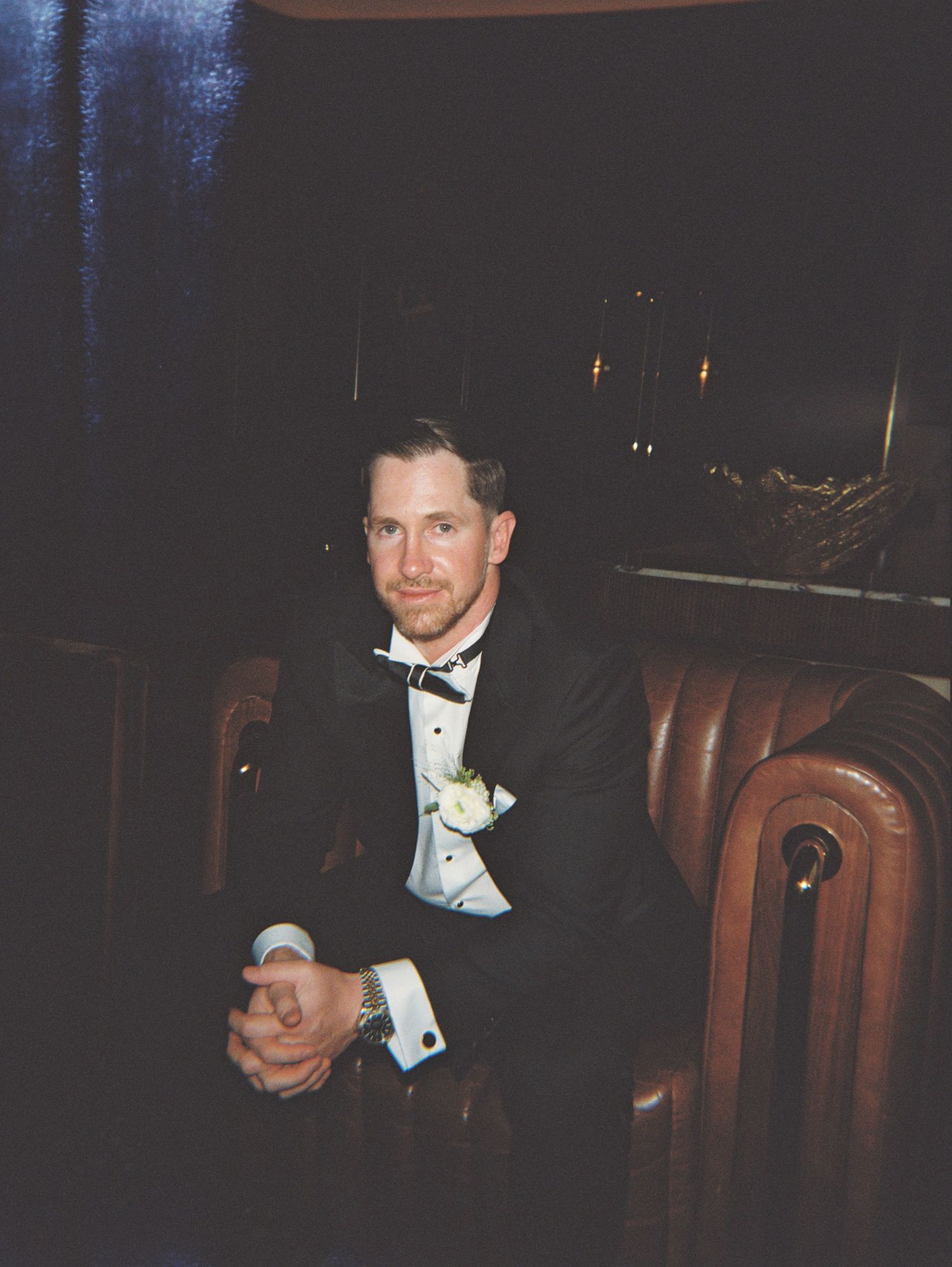 A man in a black tuxedo with bow tie and boutonniere sits in a leather chair at a formal event.