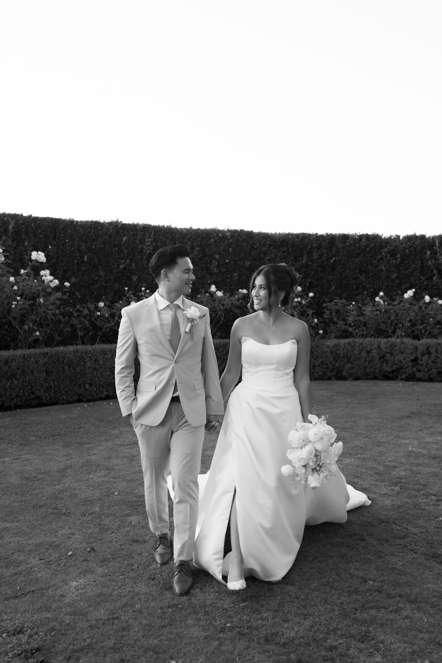 Black and white photo of a bride and groom walking hand in hand through a garden, bride holding a bouquet.