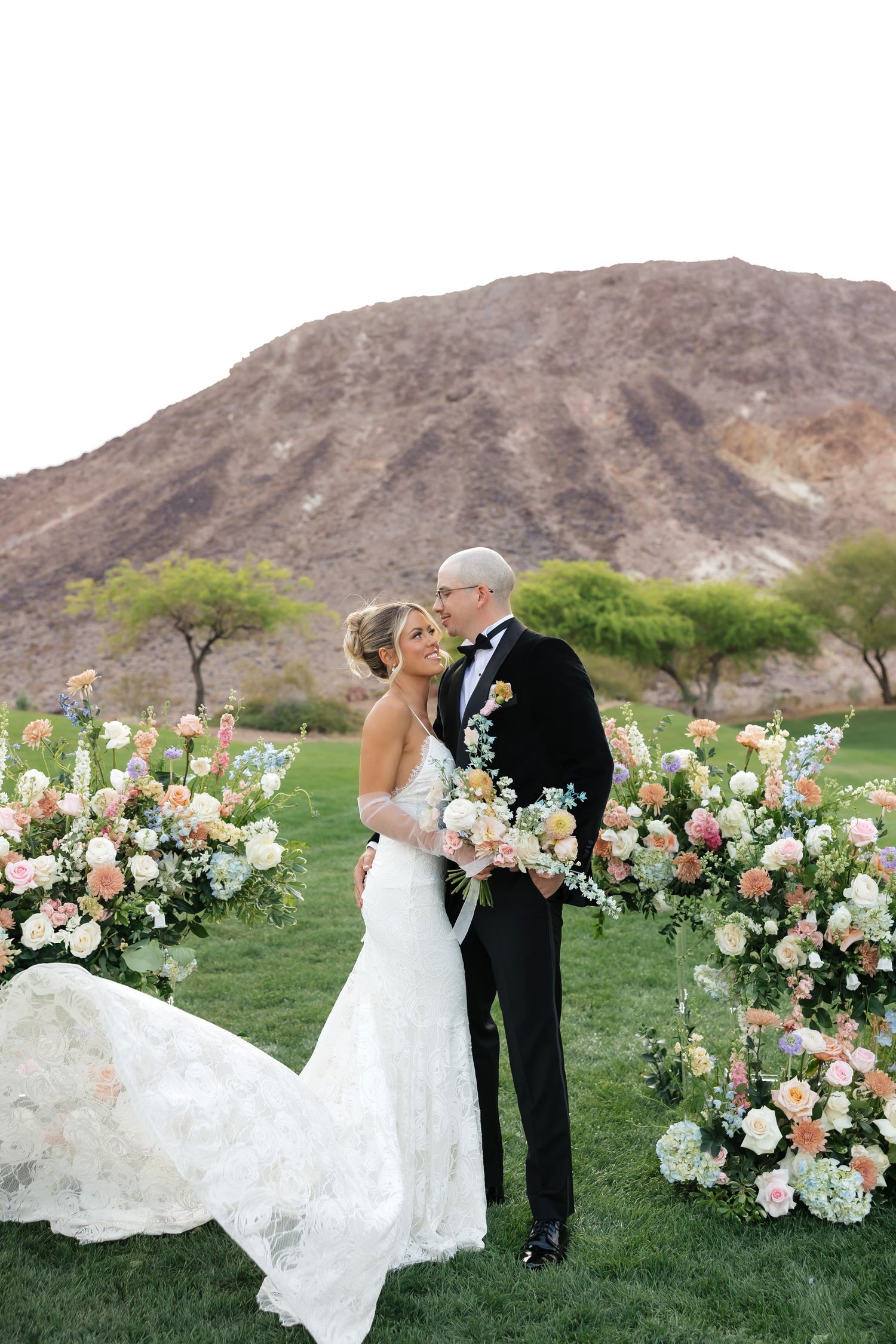 Bride and groom embrace before floral arch with desert mountain backdrop at outdoor wedding ceremony at Cascata golf course
