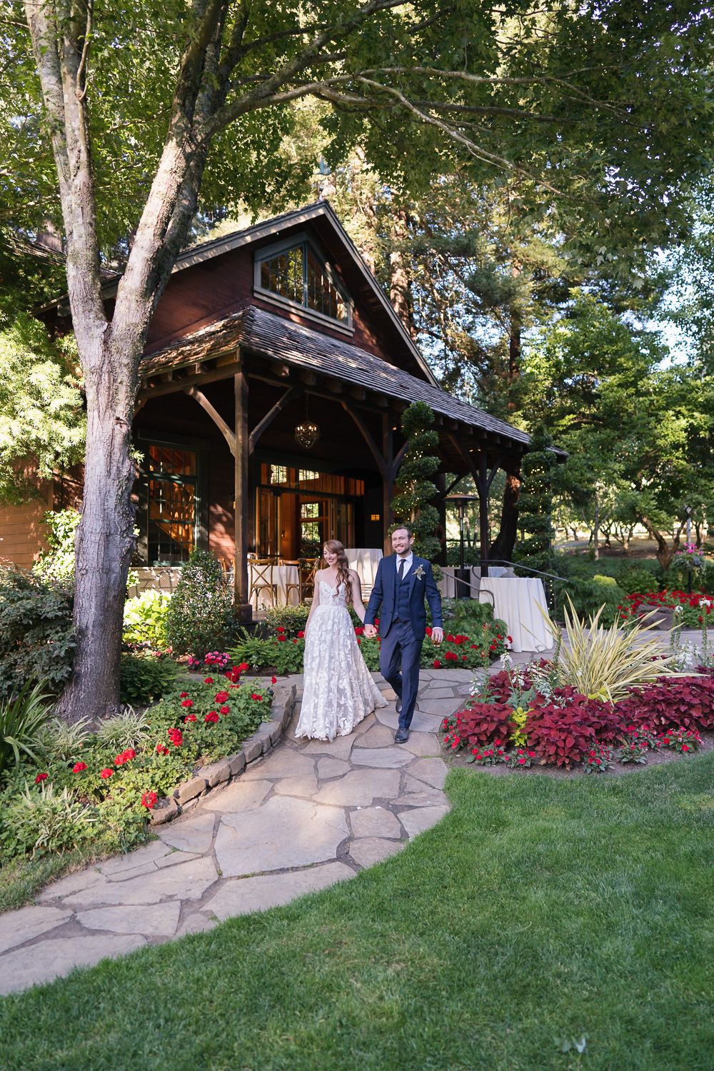 A couple walking along a stone path through beautiful gardens outside a rustic wooden chapel with a covered porch.
