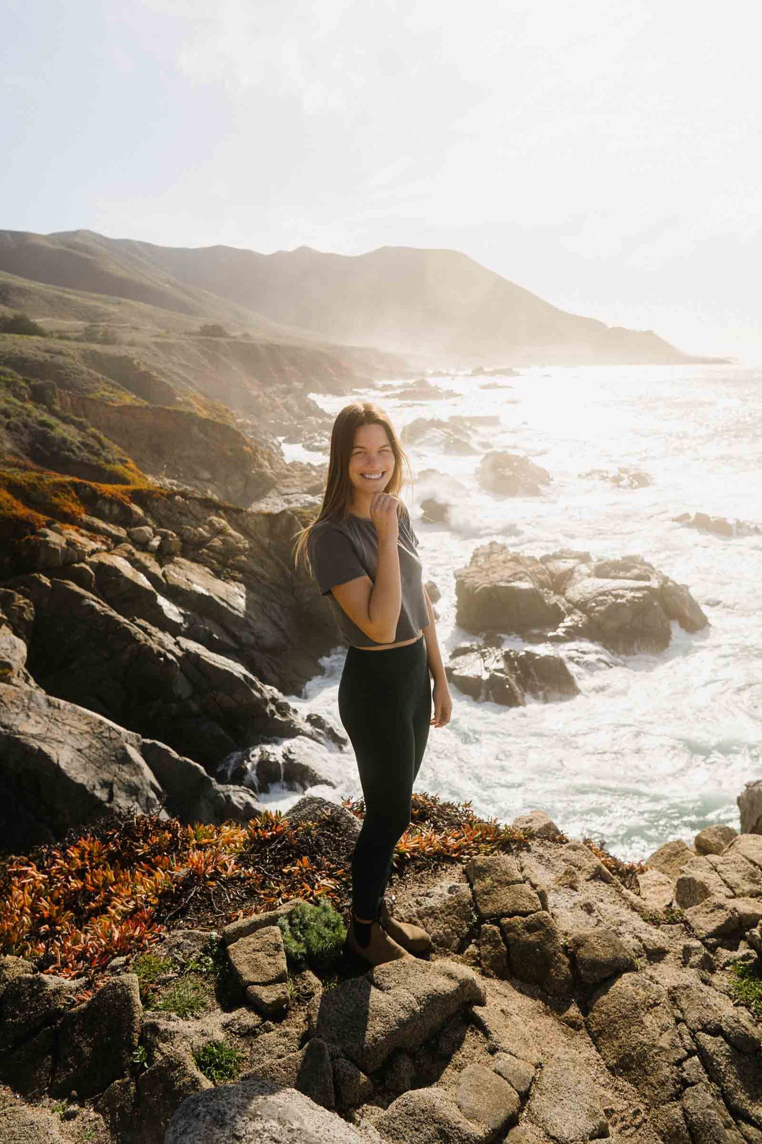 Dramatic coastal landscape with waves crashing against rocky cliffs during golden hour as figure stands on rugged terrain.