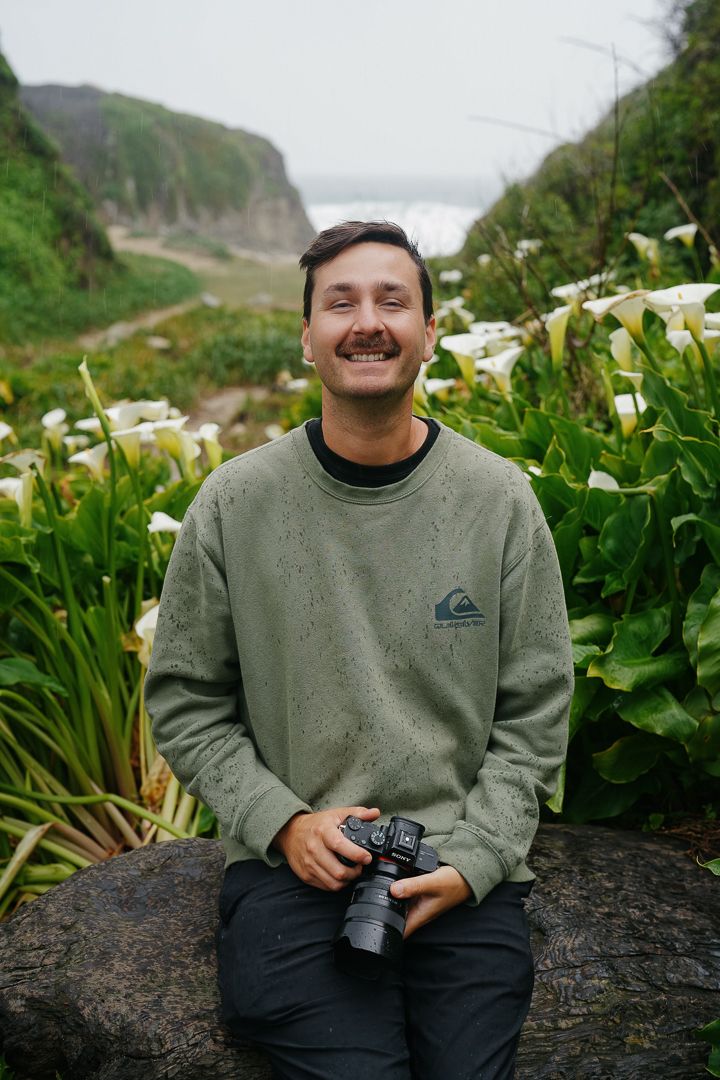 Someone wearing a gray Quiksilver sweatshirt sits on a rock with a camera amid blooming calla lilies on a coastal hillside.