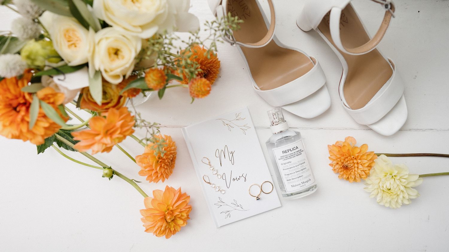 Wedding flatlay with white roses, orange dahlias, and elegant bridal shoes arranged on white surface.