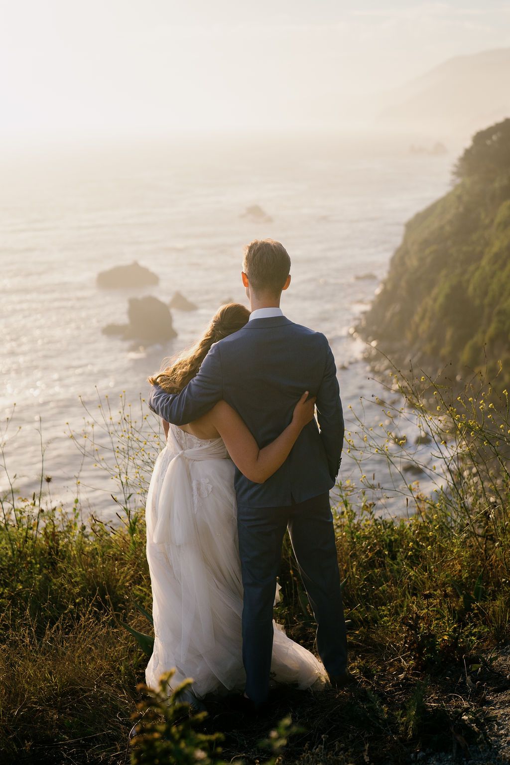 A couple embraces while overlooking a dramatic coastal cliff view at sunset along the ocean shore.