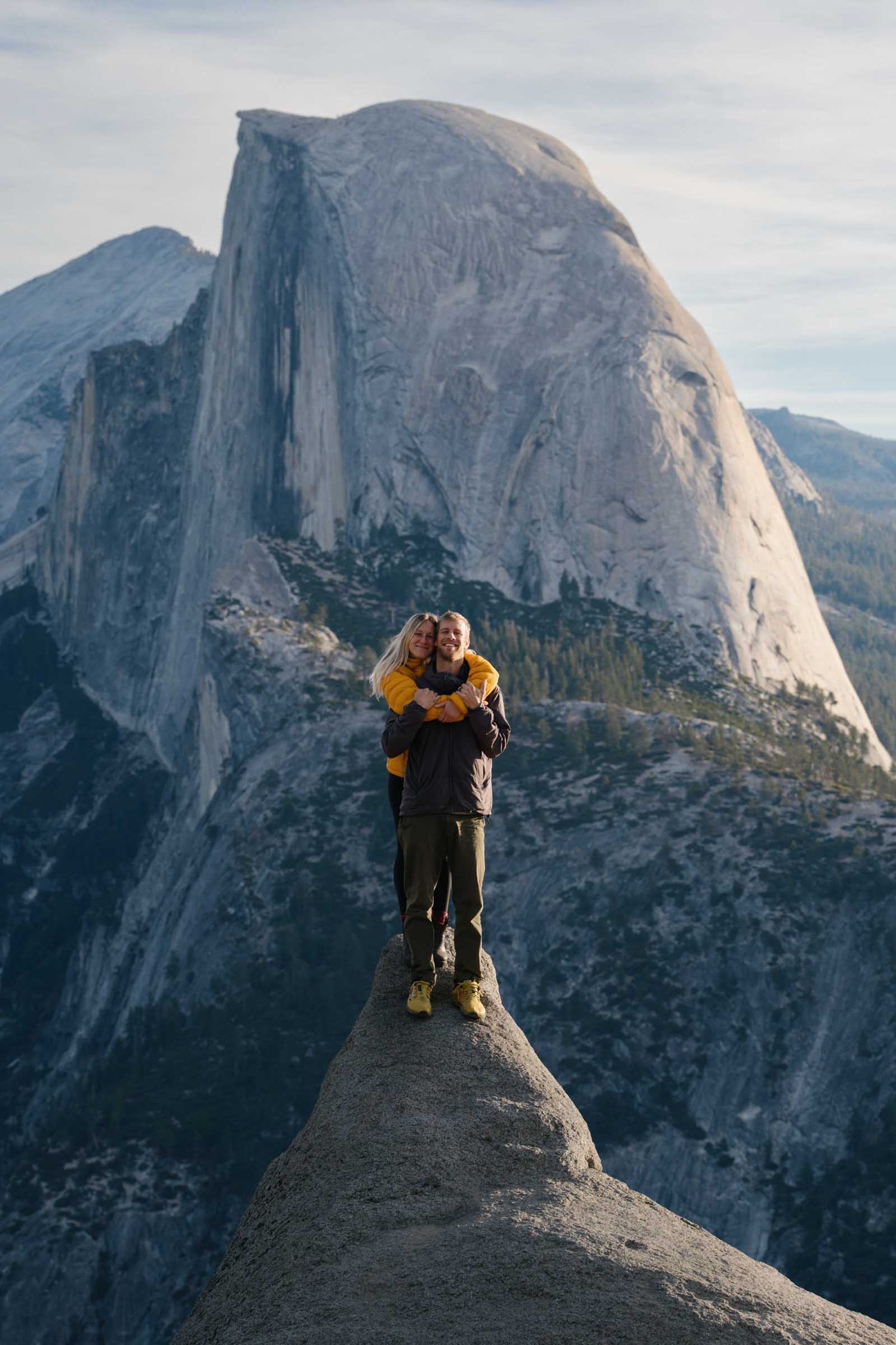Two hikers embrace while standing on the edge of Half Dome in Yosemite National Park with dramatic mountain views.