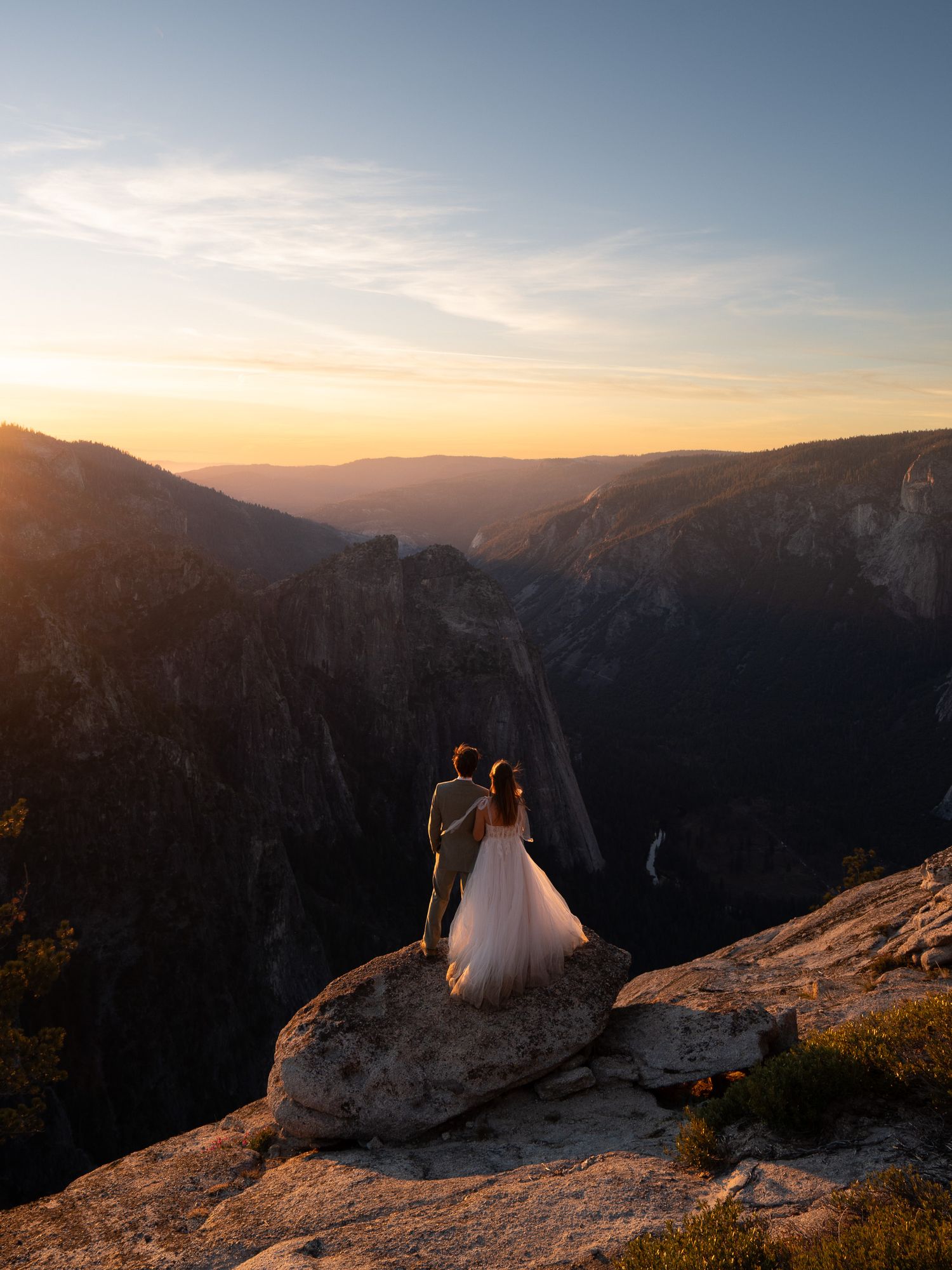 A couple sits together on a rocky mountain ledge overlooking a scenic valley at sunset in Yosemite National Park.