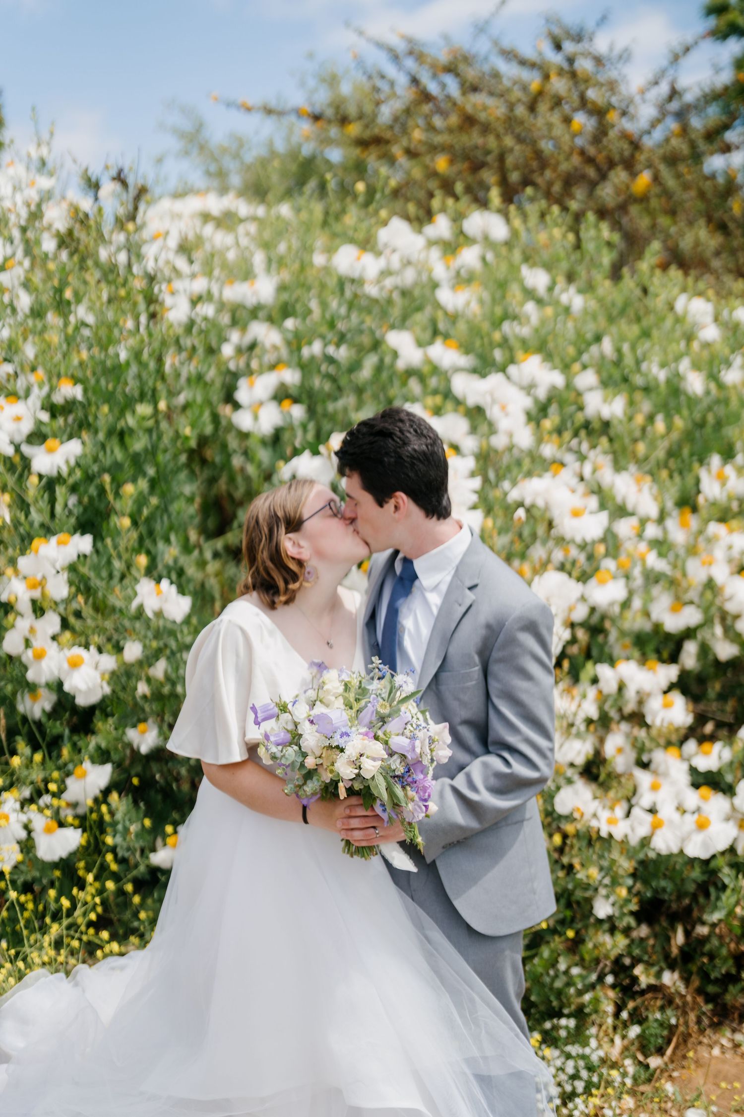 Newlyweds share a romantic kiss in a garden filled with blooming white daisies on their wedding day.