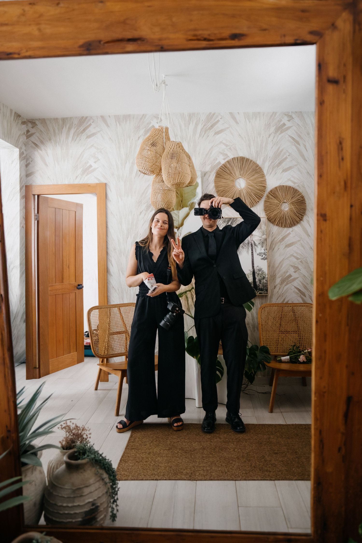 People in black clothing pose playfully with decorative fans against a white wall with palm leaf wallpaper.