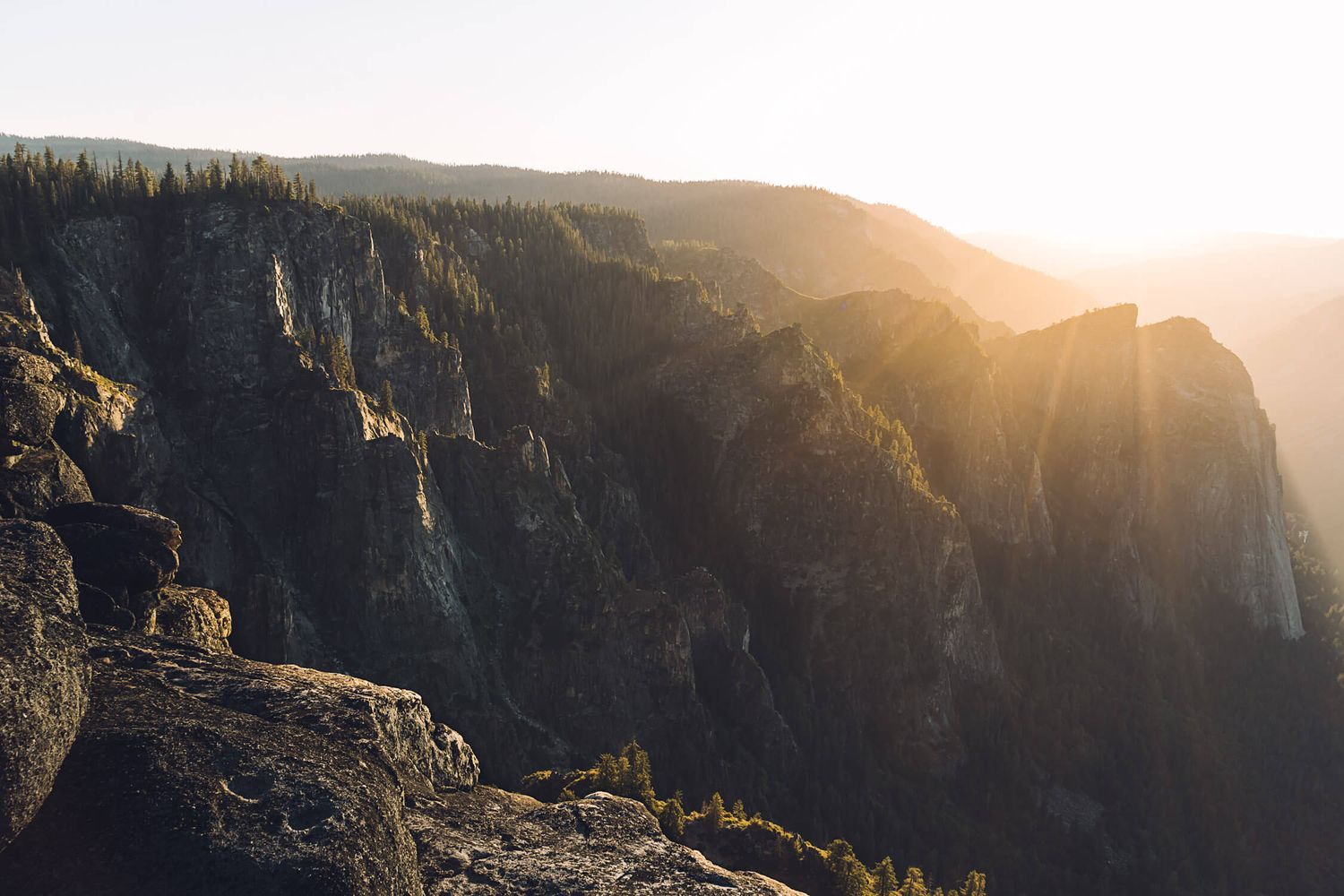 Golden sunlight streams through mountain peaks at sunset in Yosemite National Park, illuminating dramatic granite cliffs.