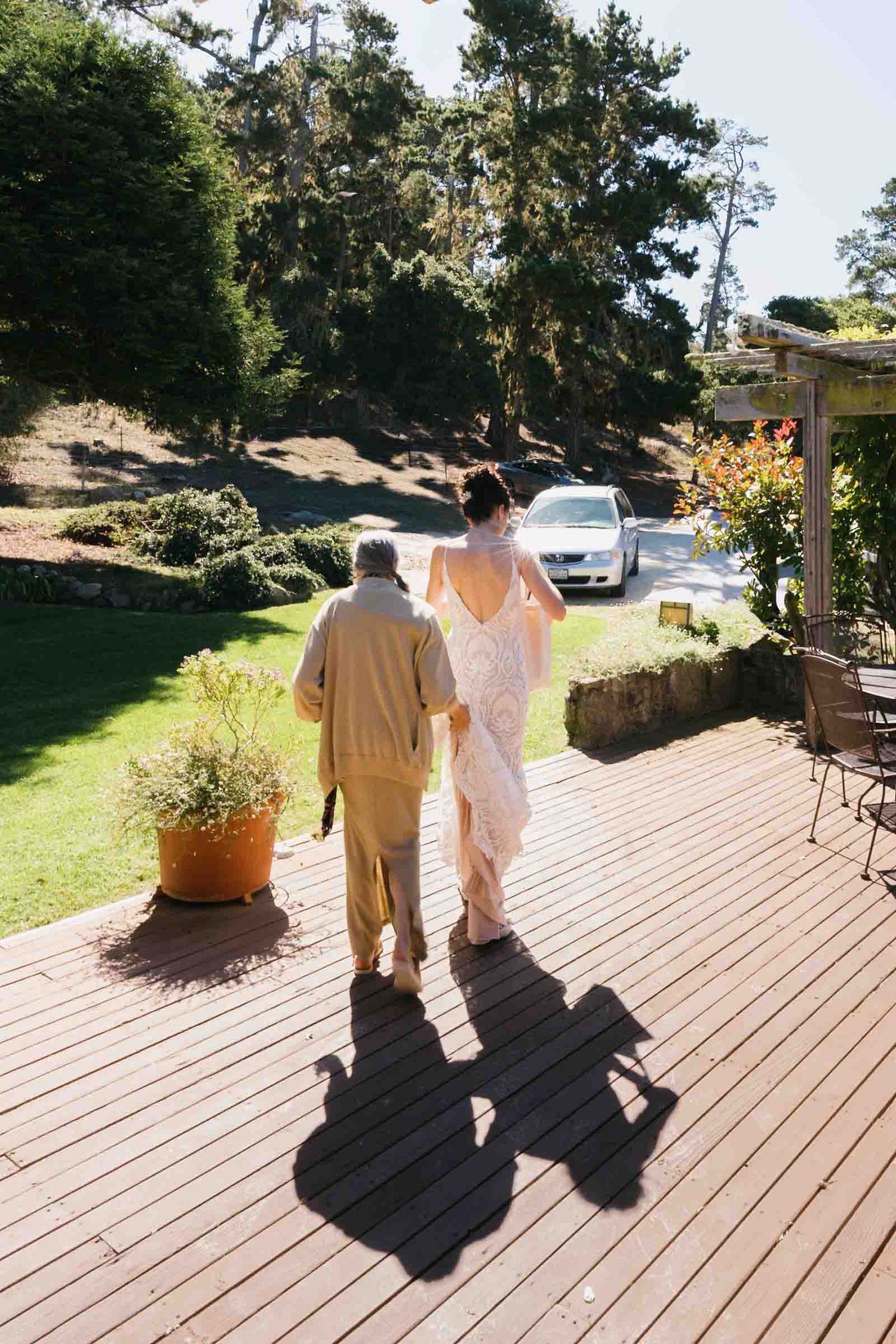 Two people walk together on a wooden deck surrounded by lush greenery on a sunny afternoon.