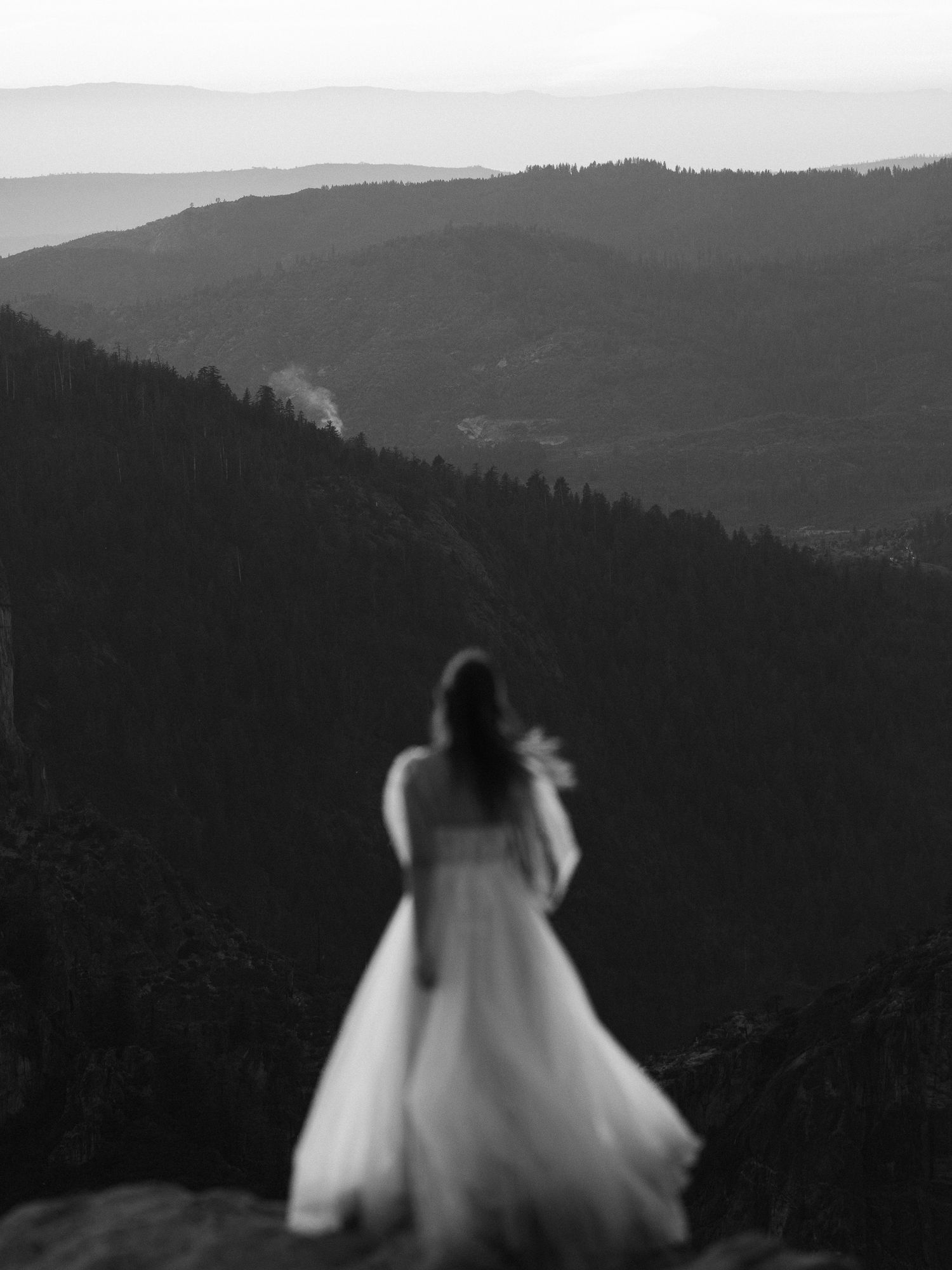 A figure in a flowing white dress stands silhouetted against a dramatic mountain landscape at sunset.