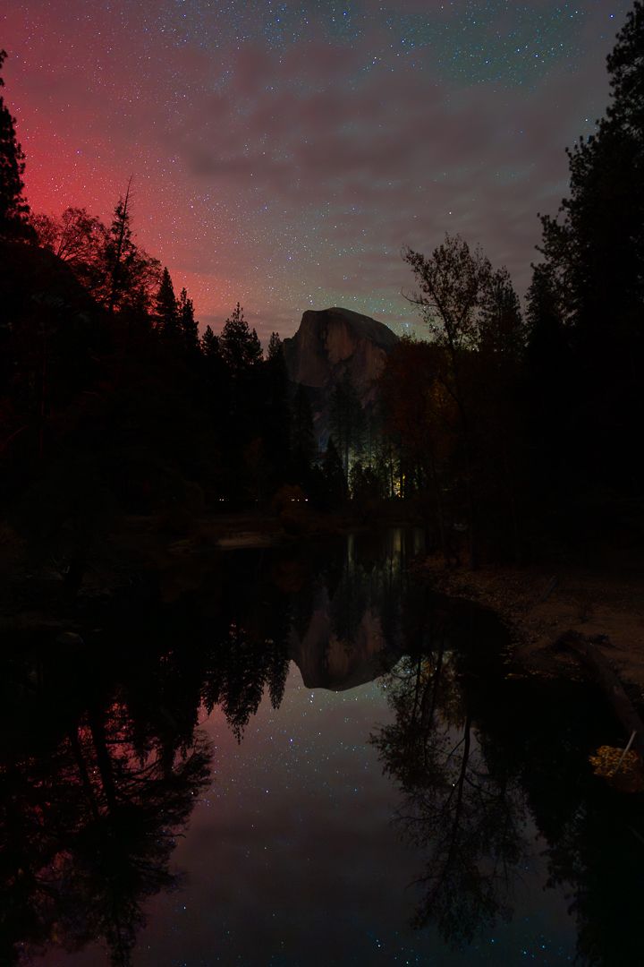 Silhouetted trees frame a tranquil lake reflecting Half Dome in Yosemite National Park under a the northern lights.