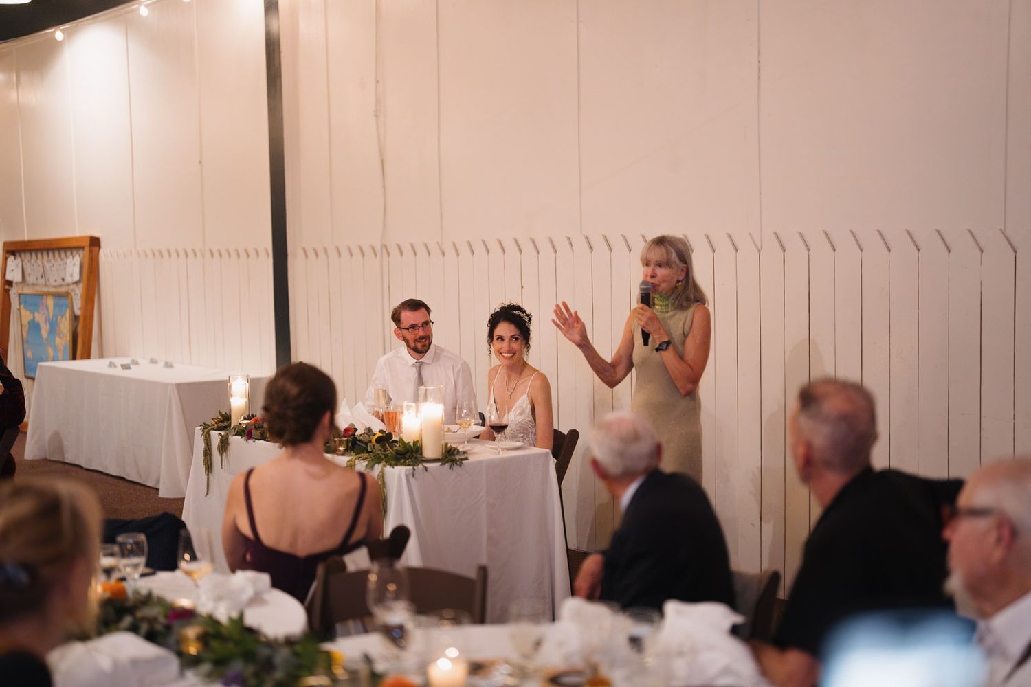 Wedding reception guests gather in a banquet hall as someone delivers a speech at the head table during dinner.