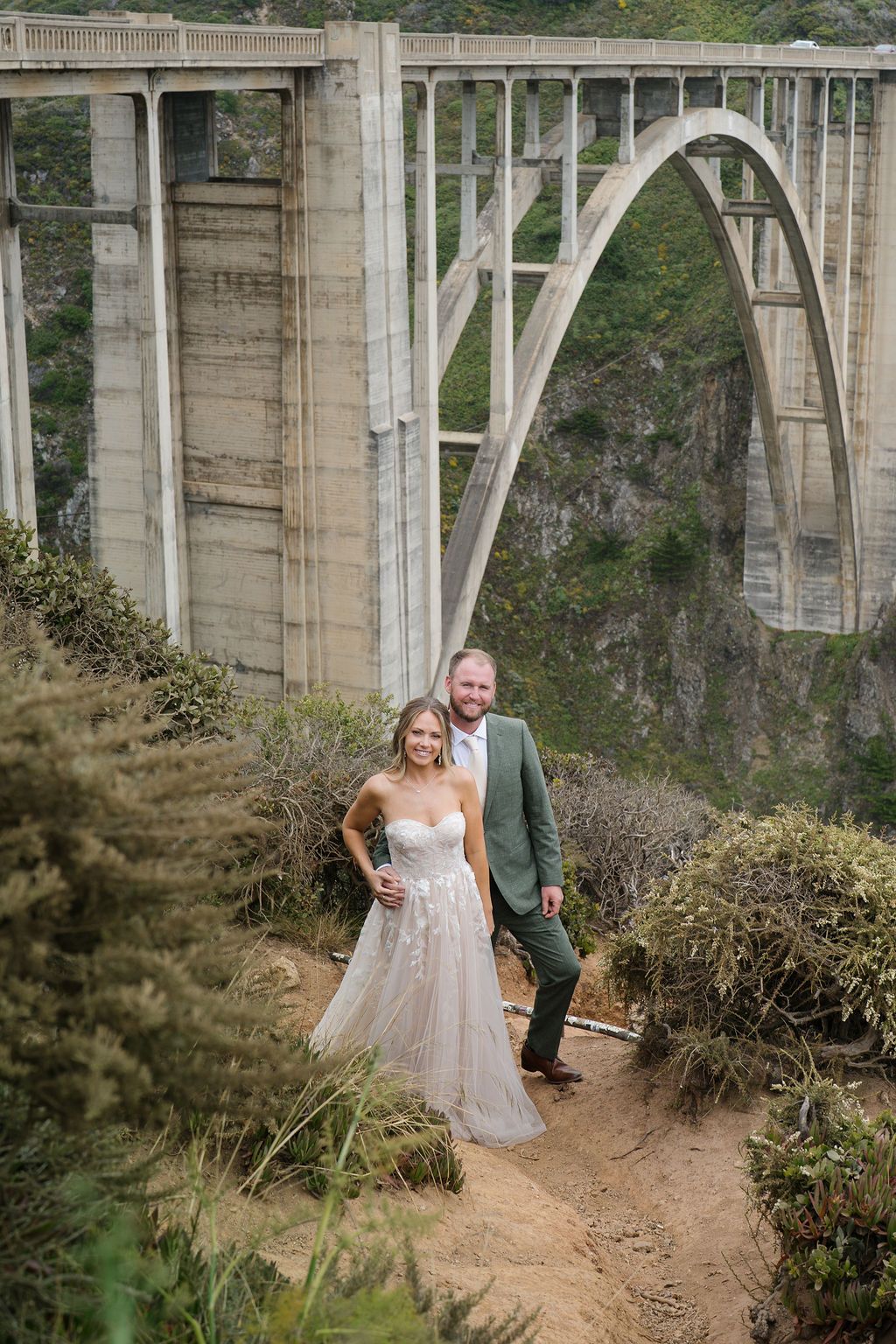 A couple poses on a hiking trail with the majestic Bixby Creek Bridge in Big Sur, California as their backdrop.