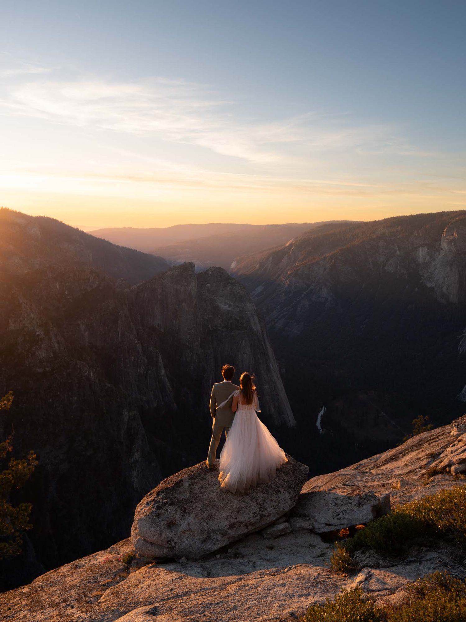 A couple in wedding attire sits on a mountain cliff edge overlooking a scenic Yosemite Valley standing on Taft Point.
