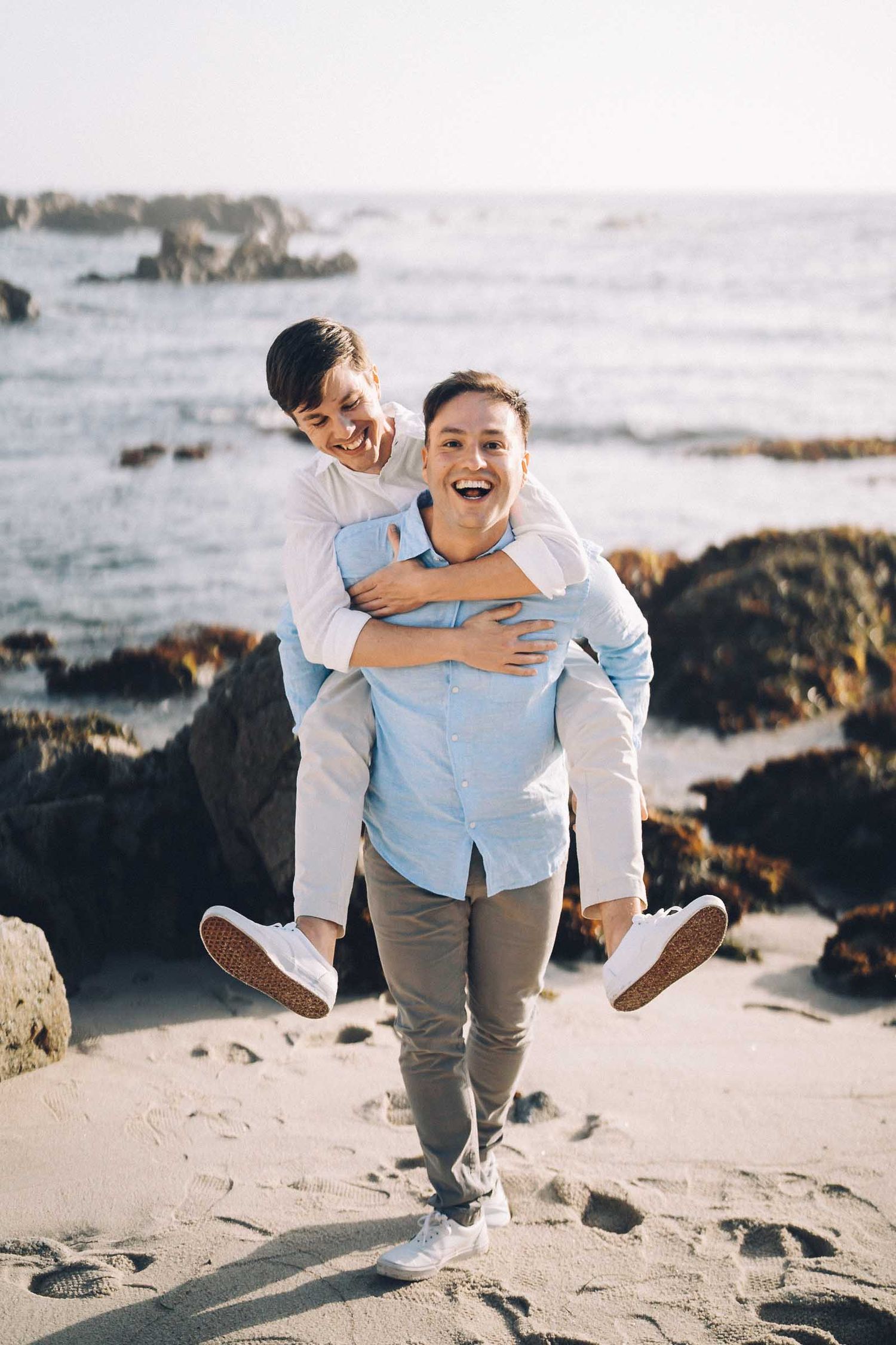 A couple shares playful moments together on a rocky beach with ocean waves and cliffs in the background.
