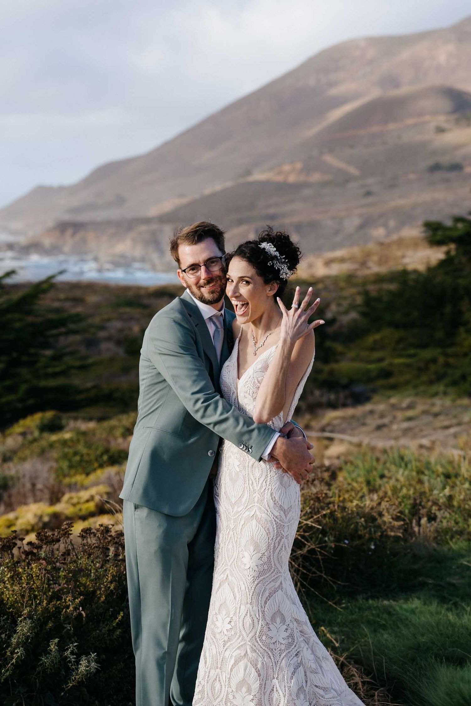 A couple shares a romantic moment against a scenic coastal mountain landscape during their wedding photoshoot.